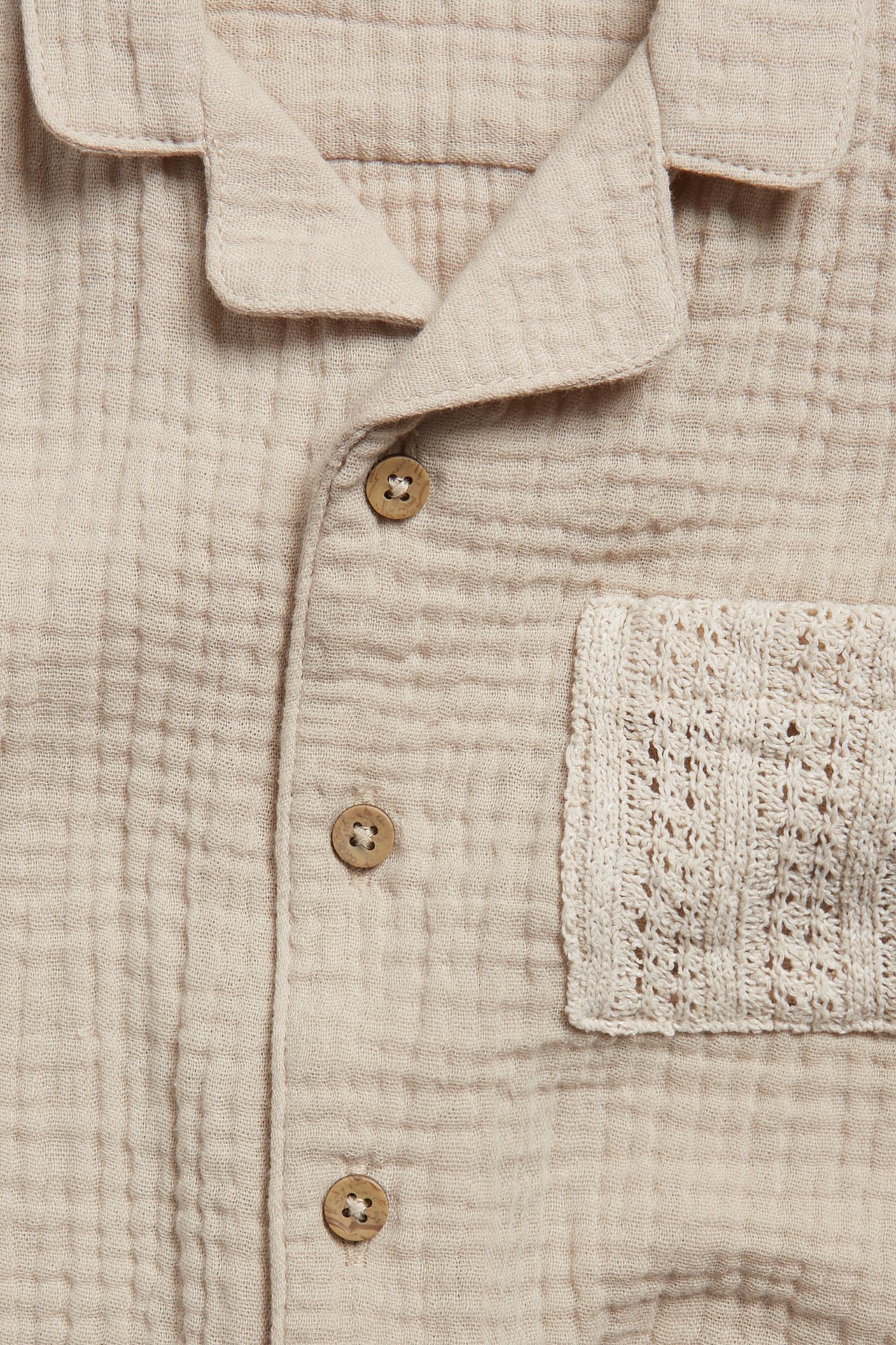 Close-up front view of a beige cotton muslin shirt with wooden buttons and a textured knit chest pocket.