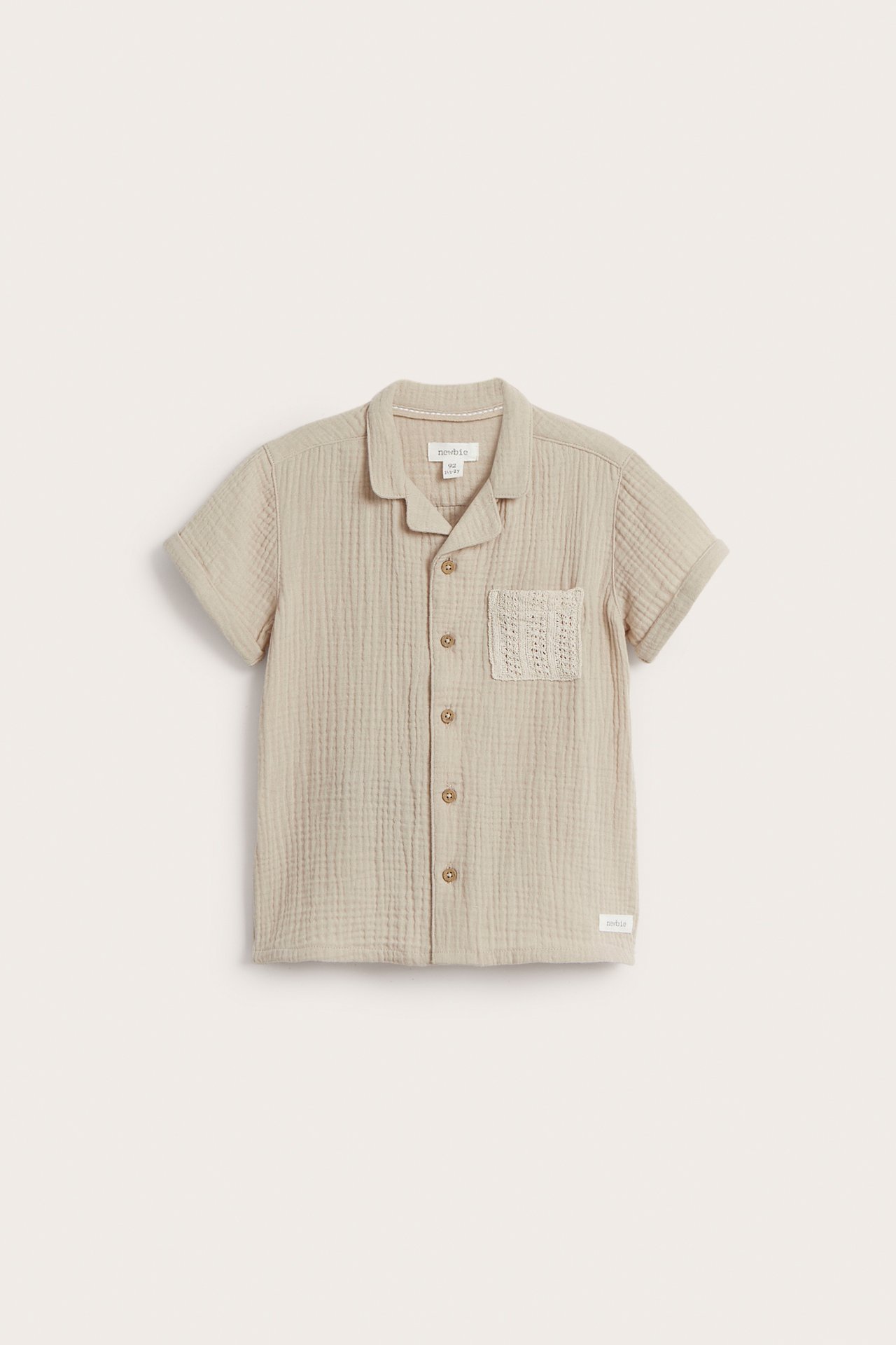 Front view of a kid's beige cotton muslin short-sleeved shirt with a knitted chest pocket and button-front.