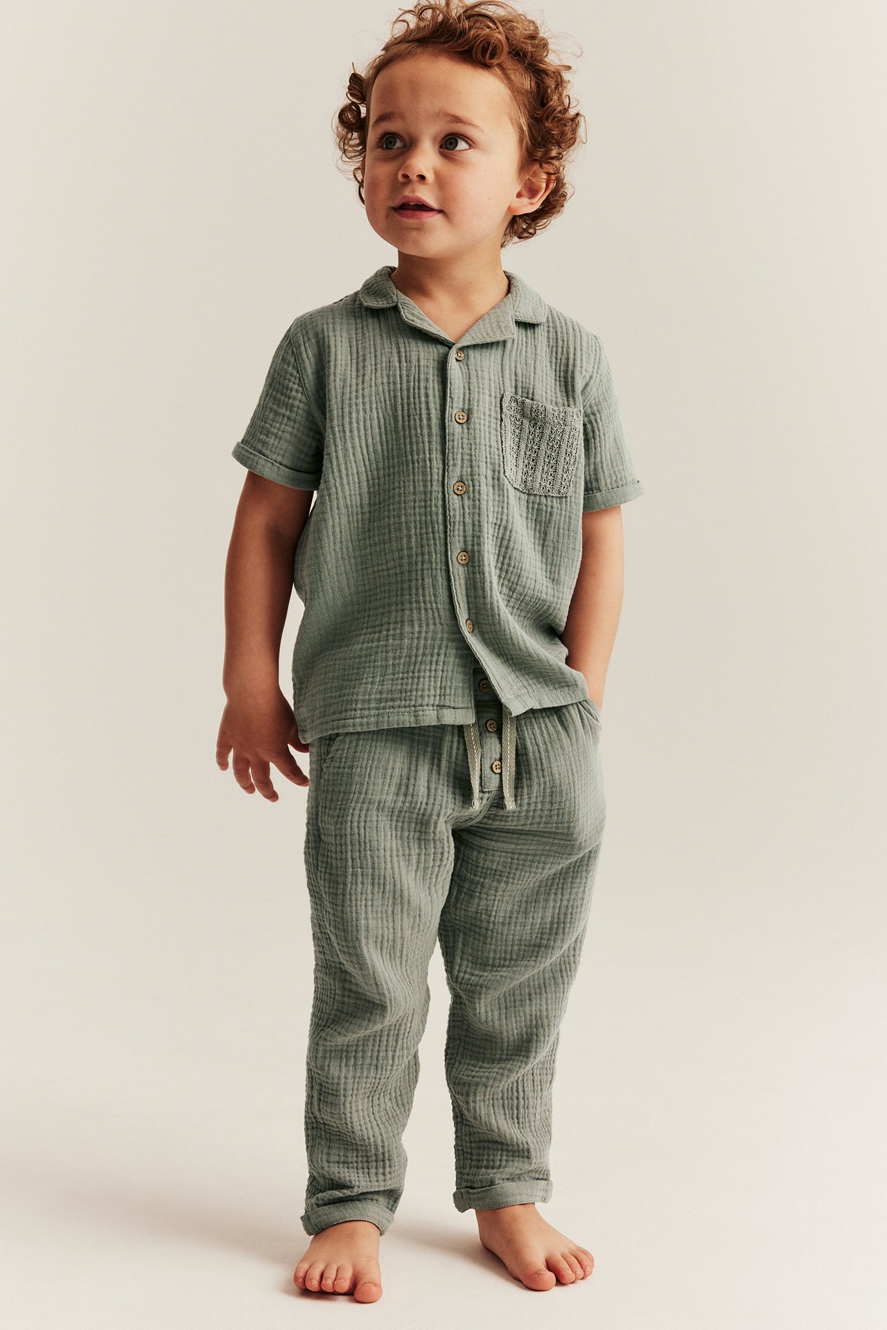 Front view: Kid in dusty green cotton muslin short-sleeved shirt with pocket and matching pants.