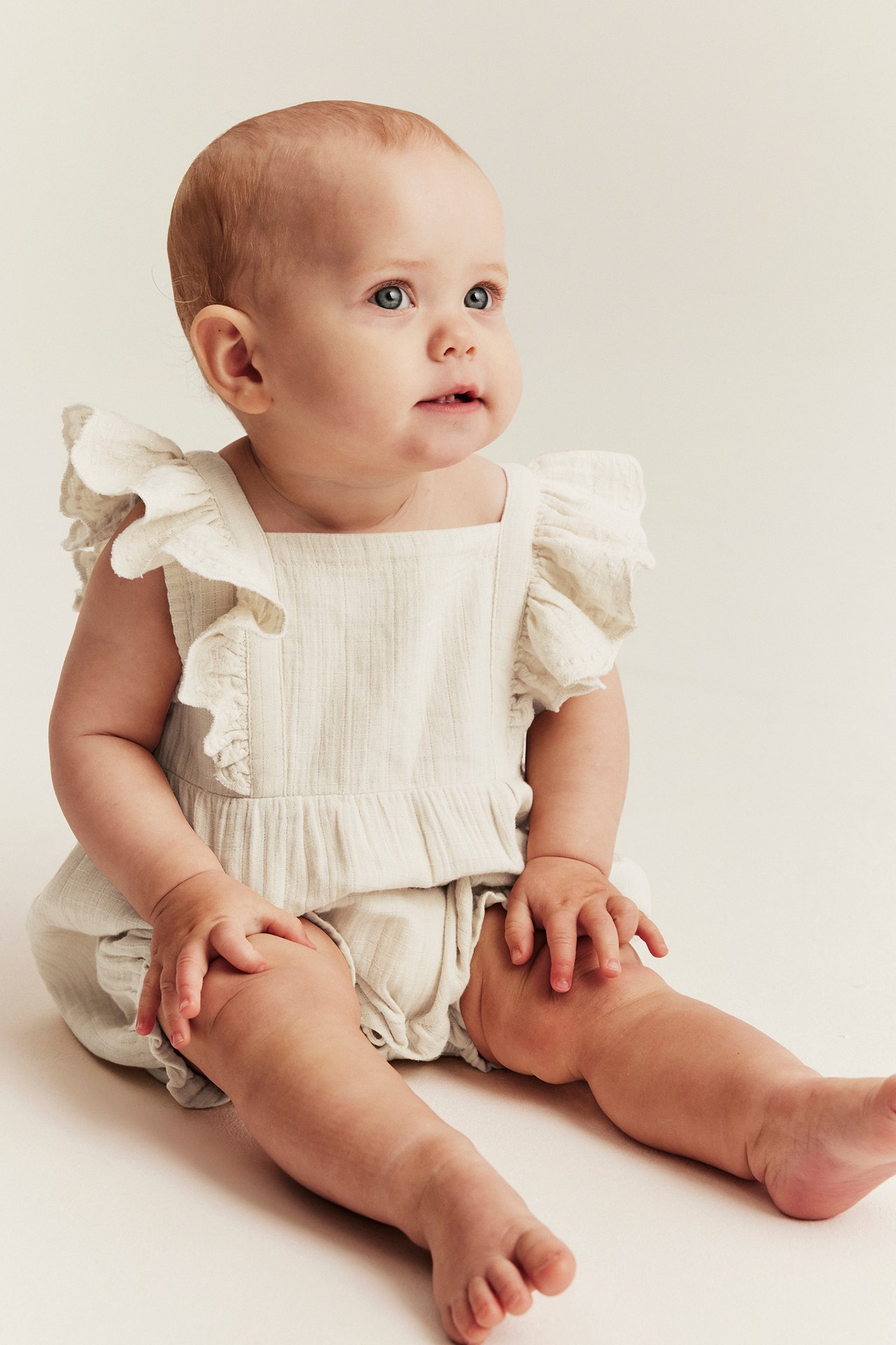 Front view of a sitting baby wearing a light beige woven cotton playsuit with ruffled straps and textured fabric.