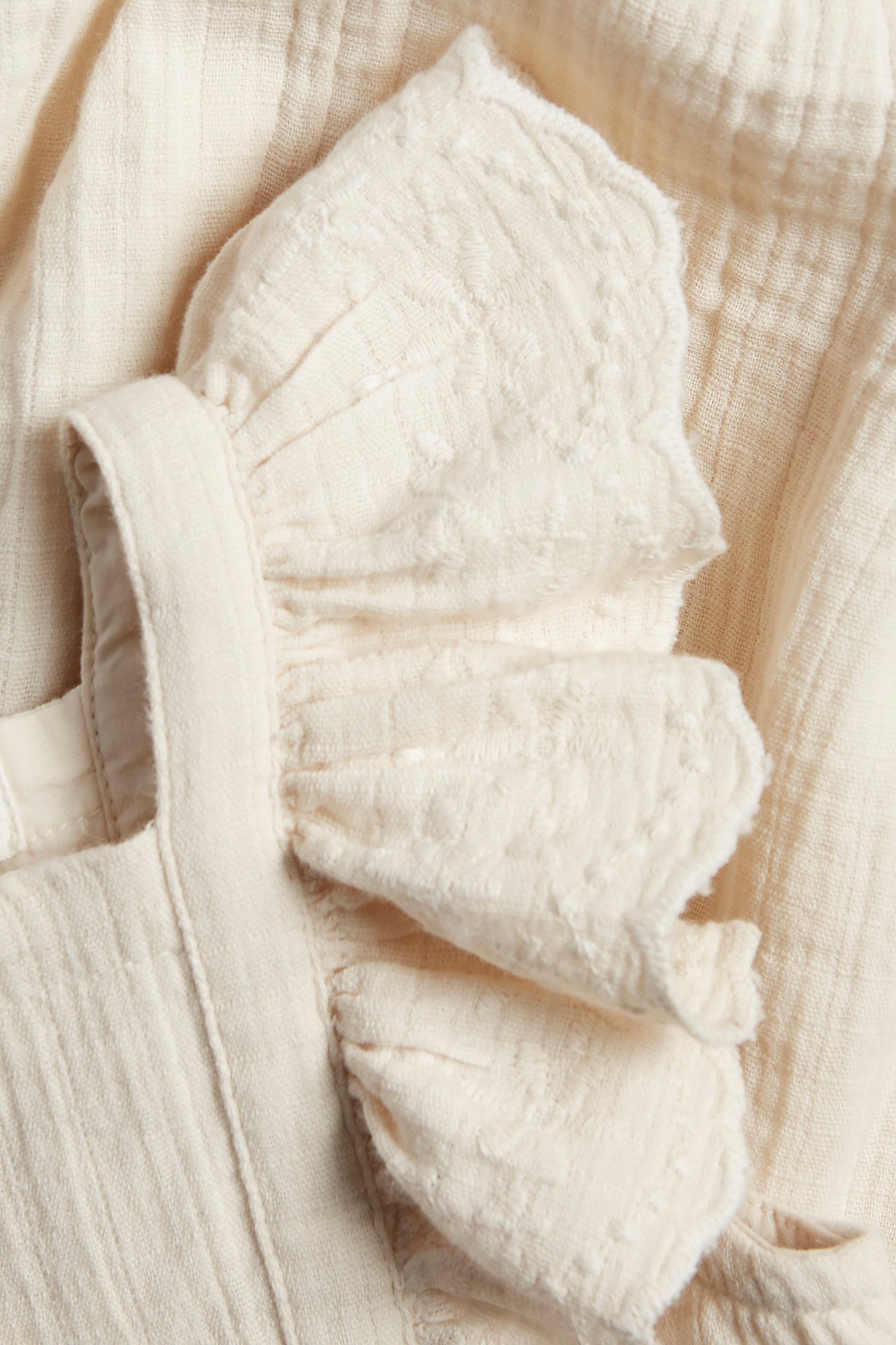 Close-up of a light beige baby cotton playsuit with textured fabric and embroidered ruffles.
