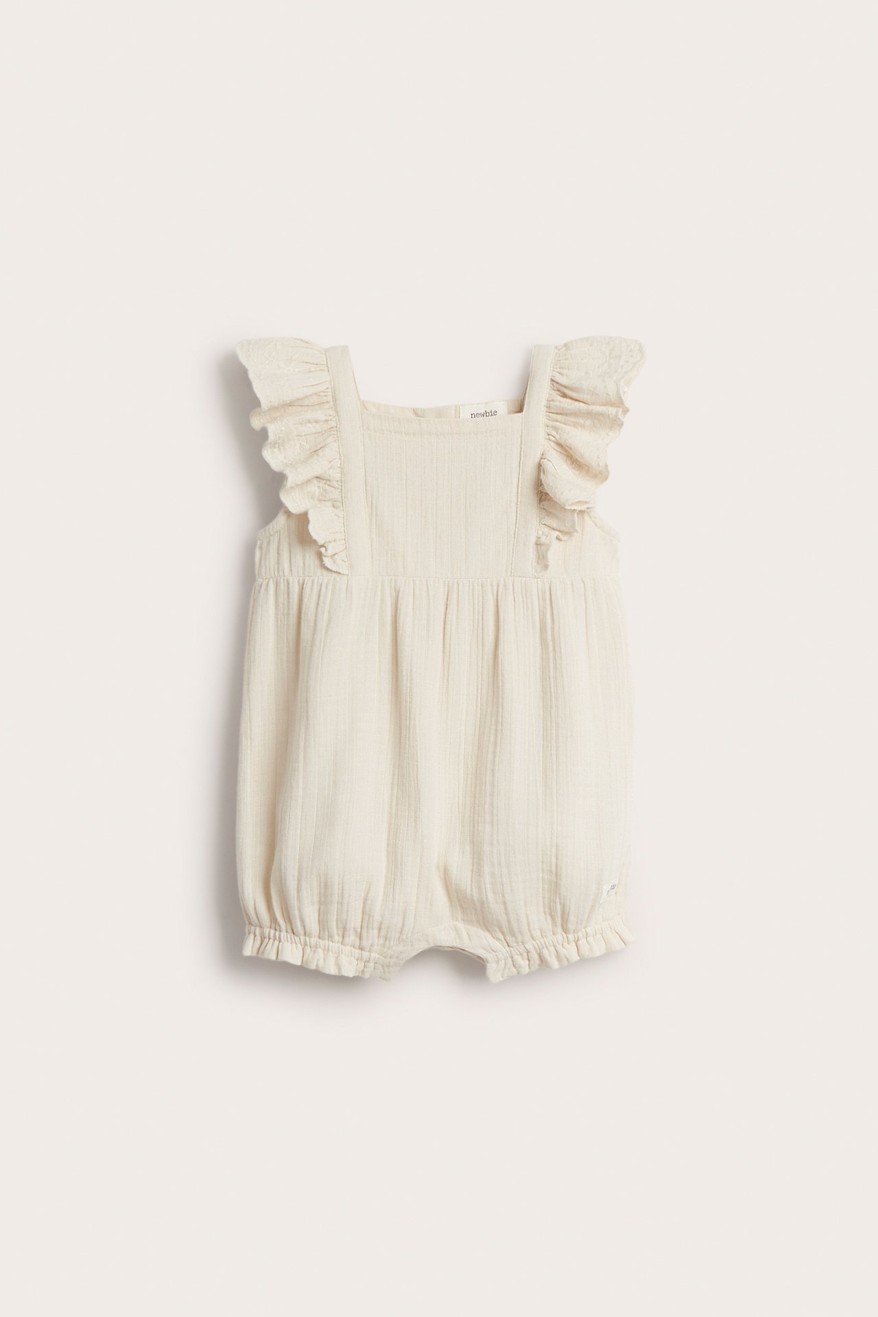 Front view of a light beige woven cotton baby playsuit with ruffled embroidered shoulders and elastic leg openings.