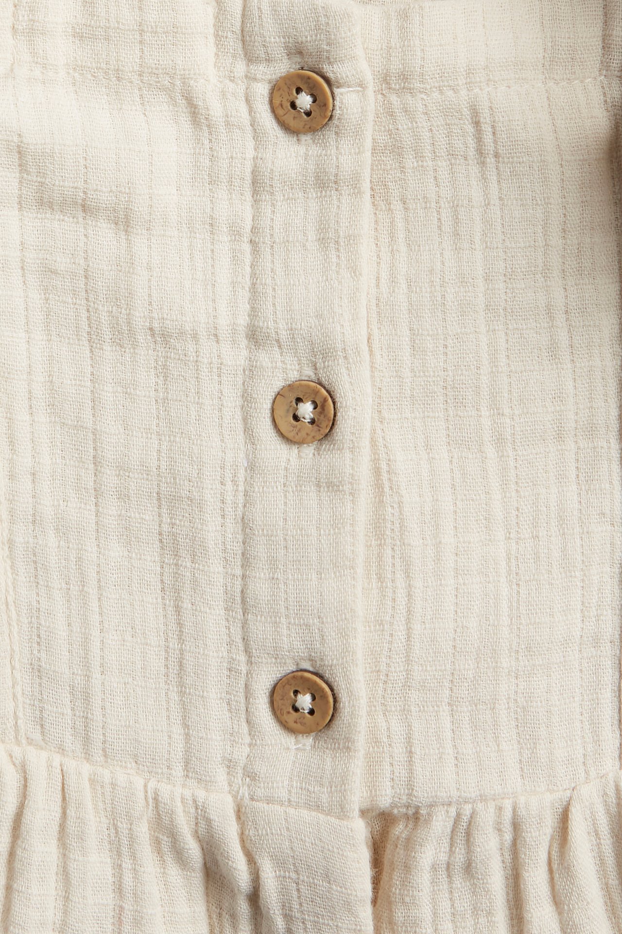 Close-up of a light beige woven cotton romper with textured fabric and three round wooden buttons.