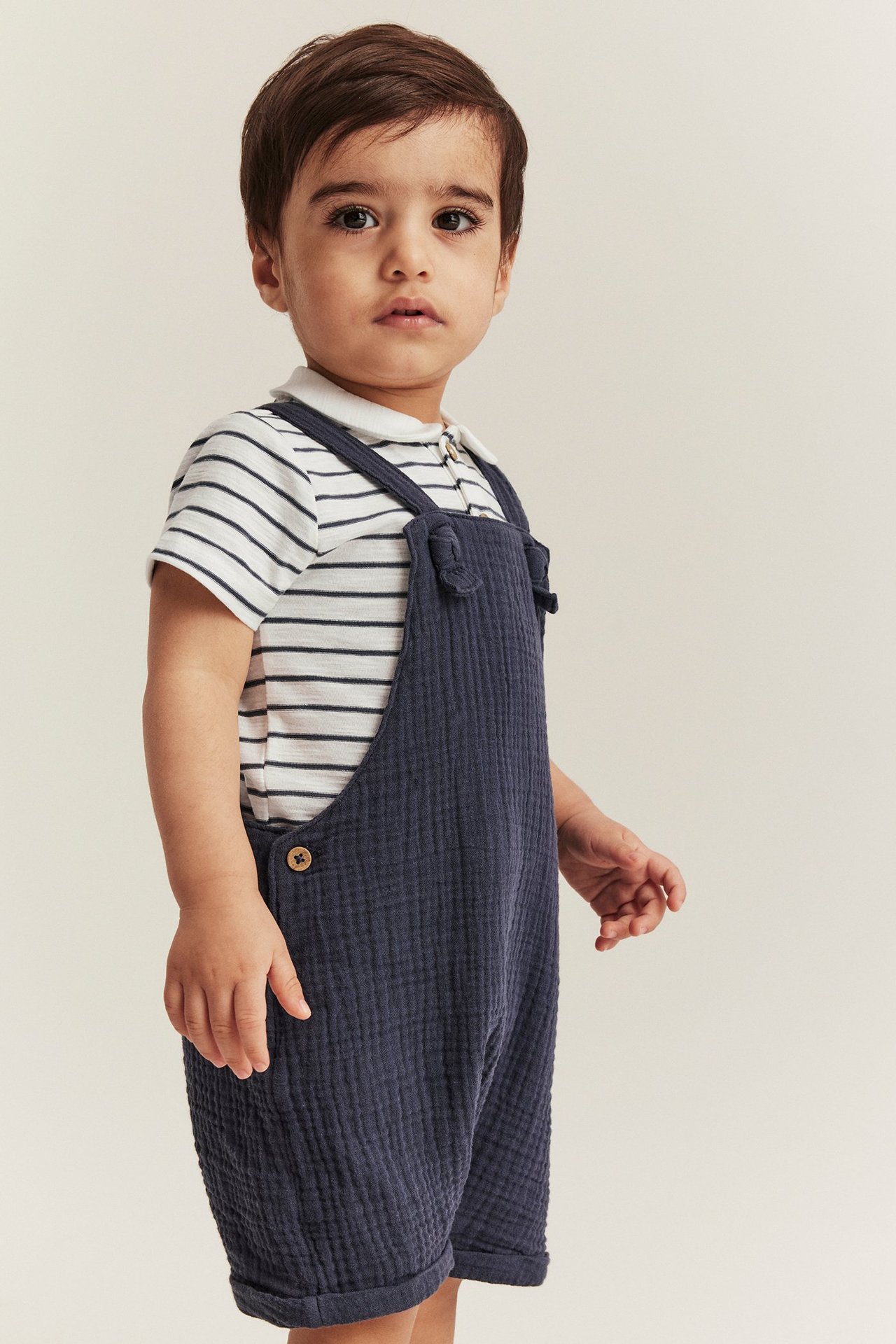 Front view of a baby wearing an off-white striped short-sleeved body with a collar, under a dark blue textured overall.