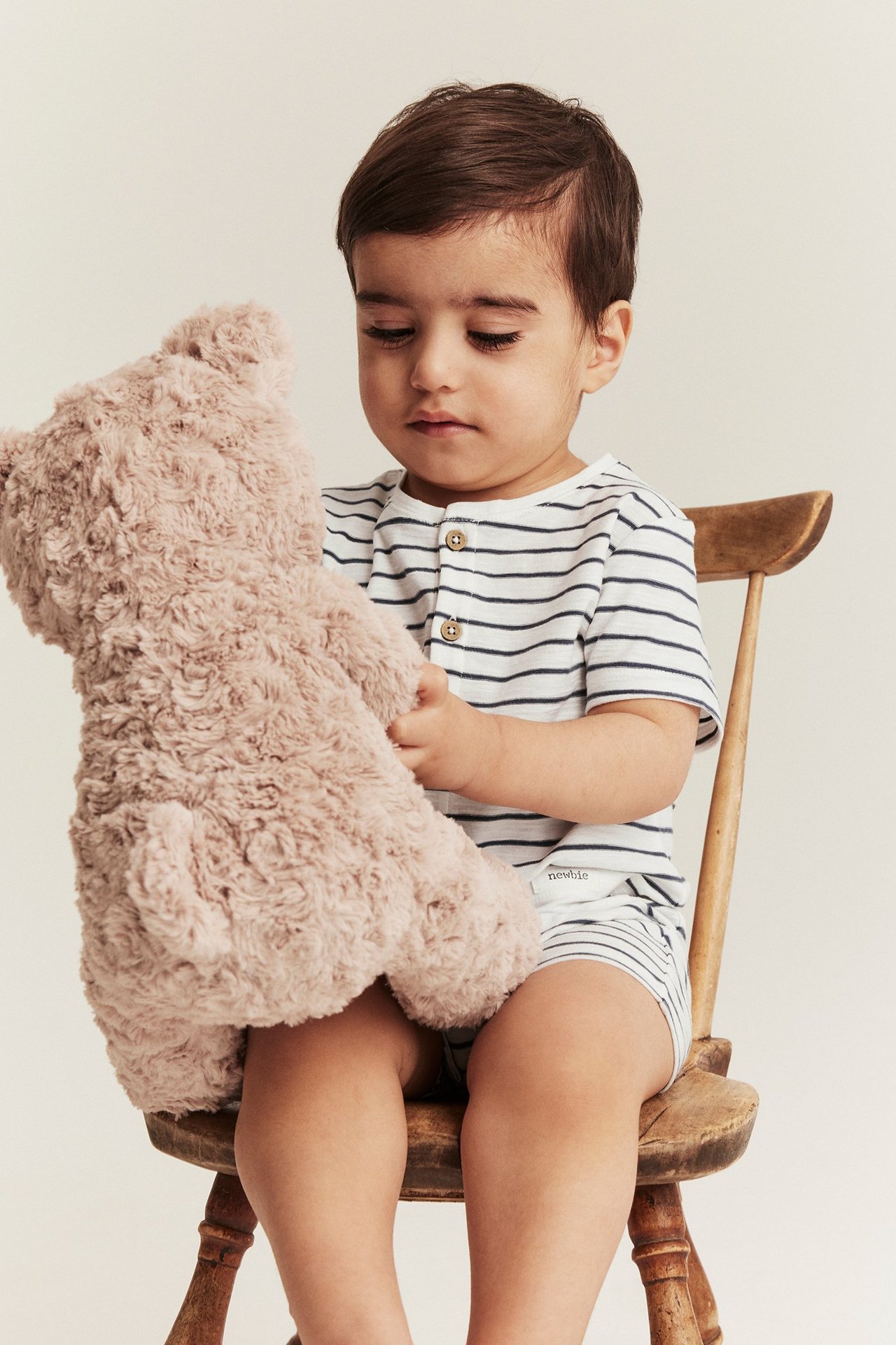 Front view of a baby wearing an off-white striped short-sleeved romper, holding a fluffy beige teddy bear.