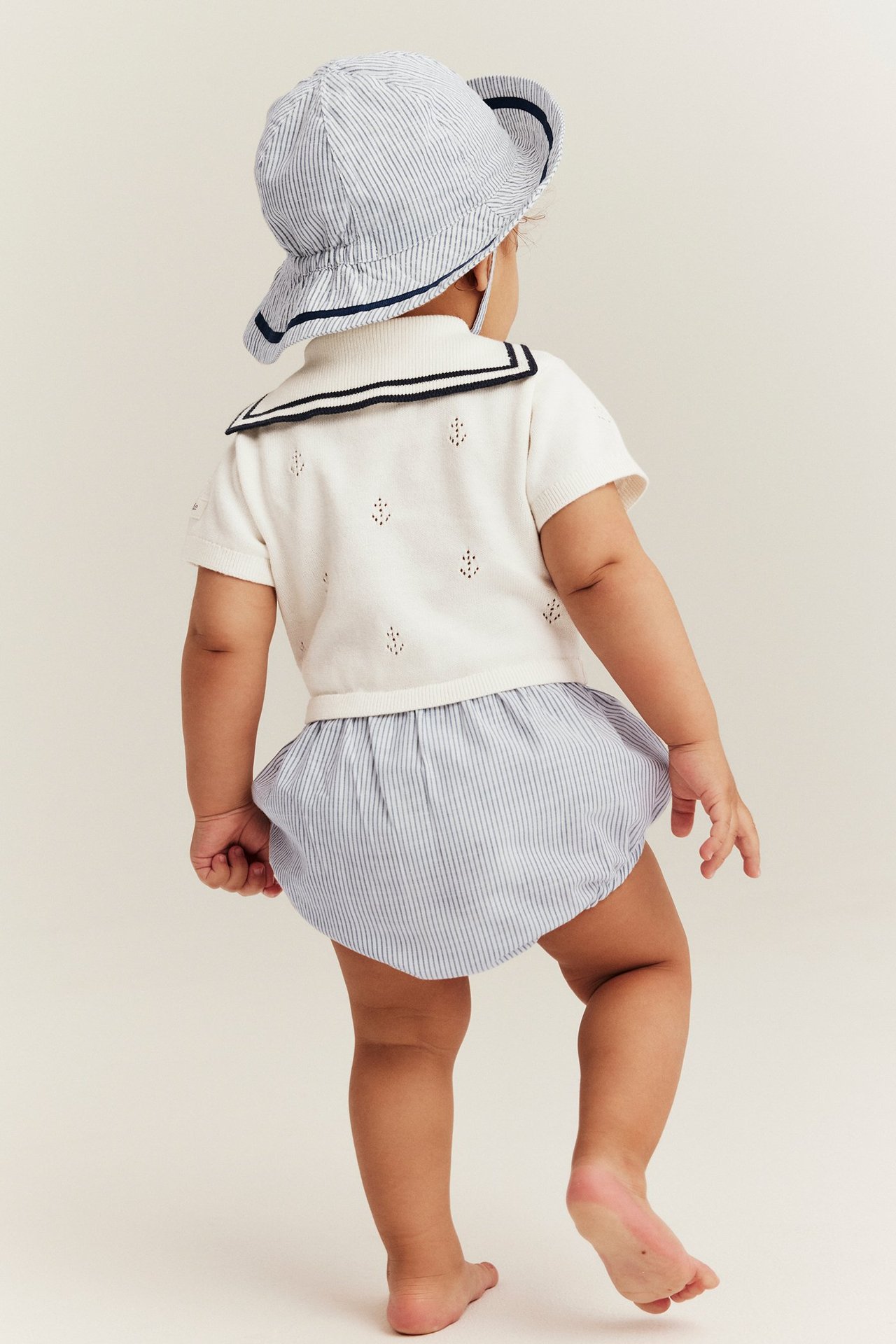Rückenansicht eines Babys in einem cremefarbenen Anker-Playsuit mit Matrosenkragen und gestreiftem Hut.