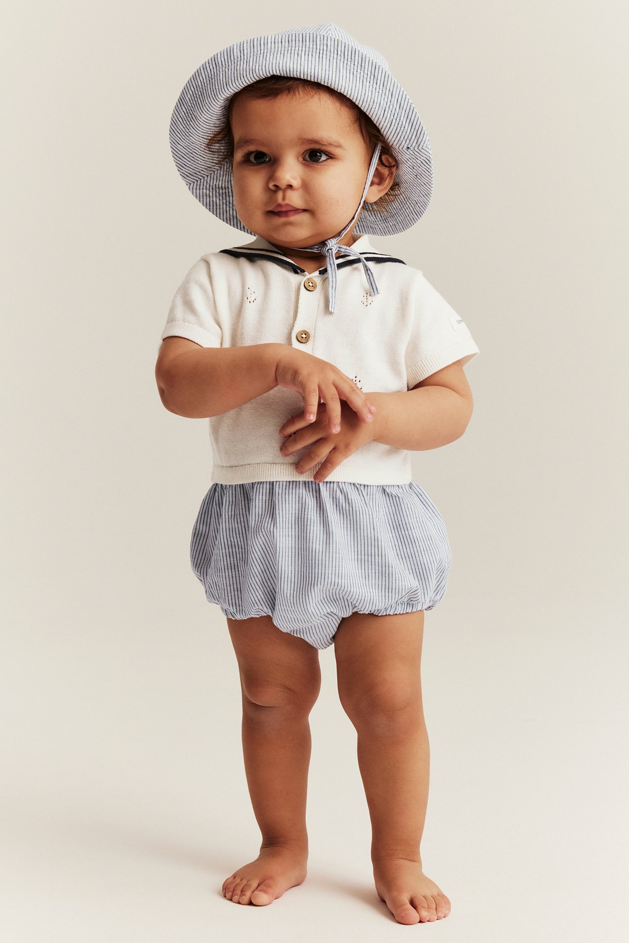 Baby trägt einen cremefarbenen Strick-Playsuit mit Matrosenkragen, gestreiftem Unterteil und gestreiften Hut.