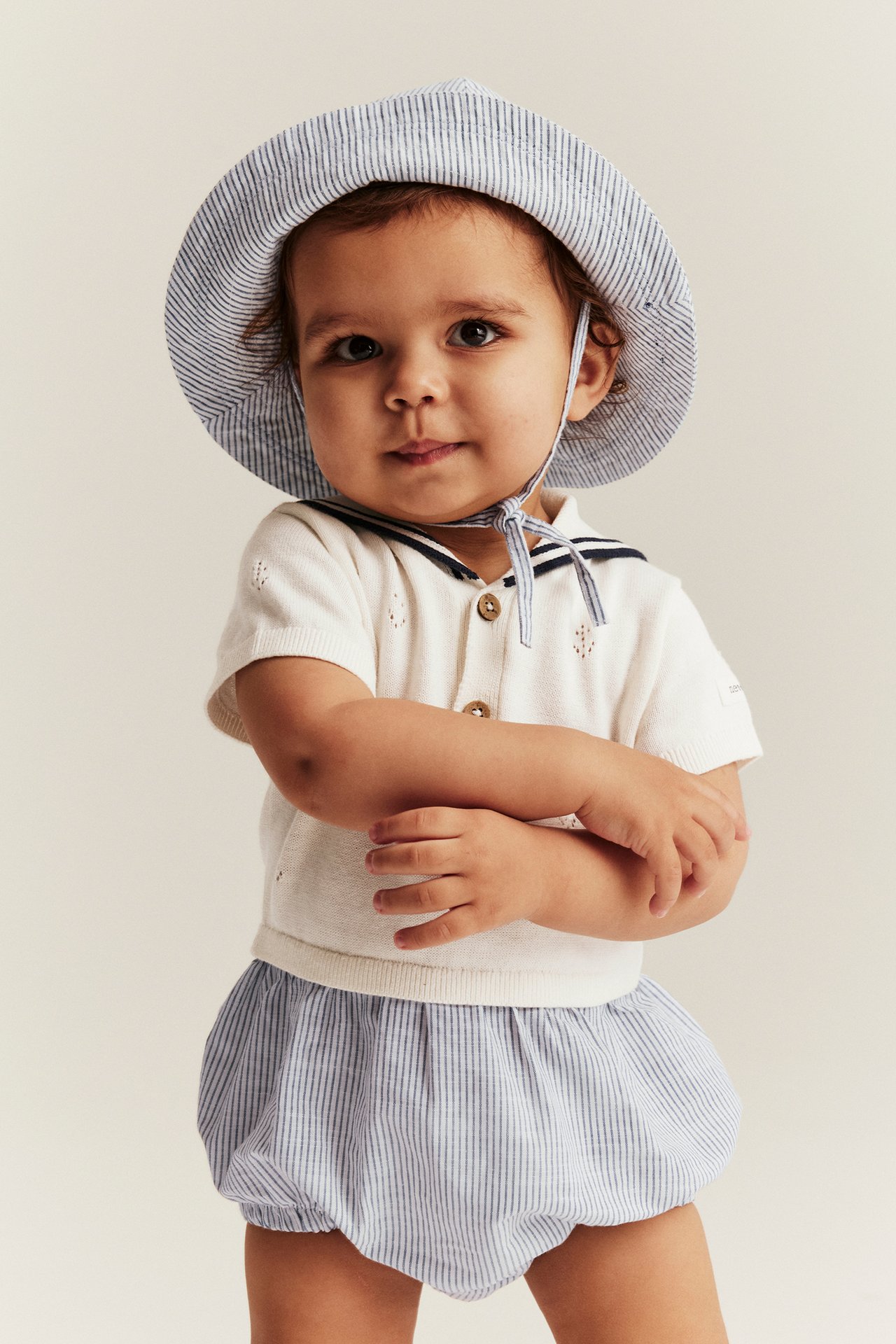 Vorderansicht eines Babys in einem cremefarbenen Strick-Strampler mit Matrosenkragen, gestreiftem Unterteil und gestreiftem Hut.