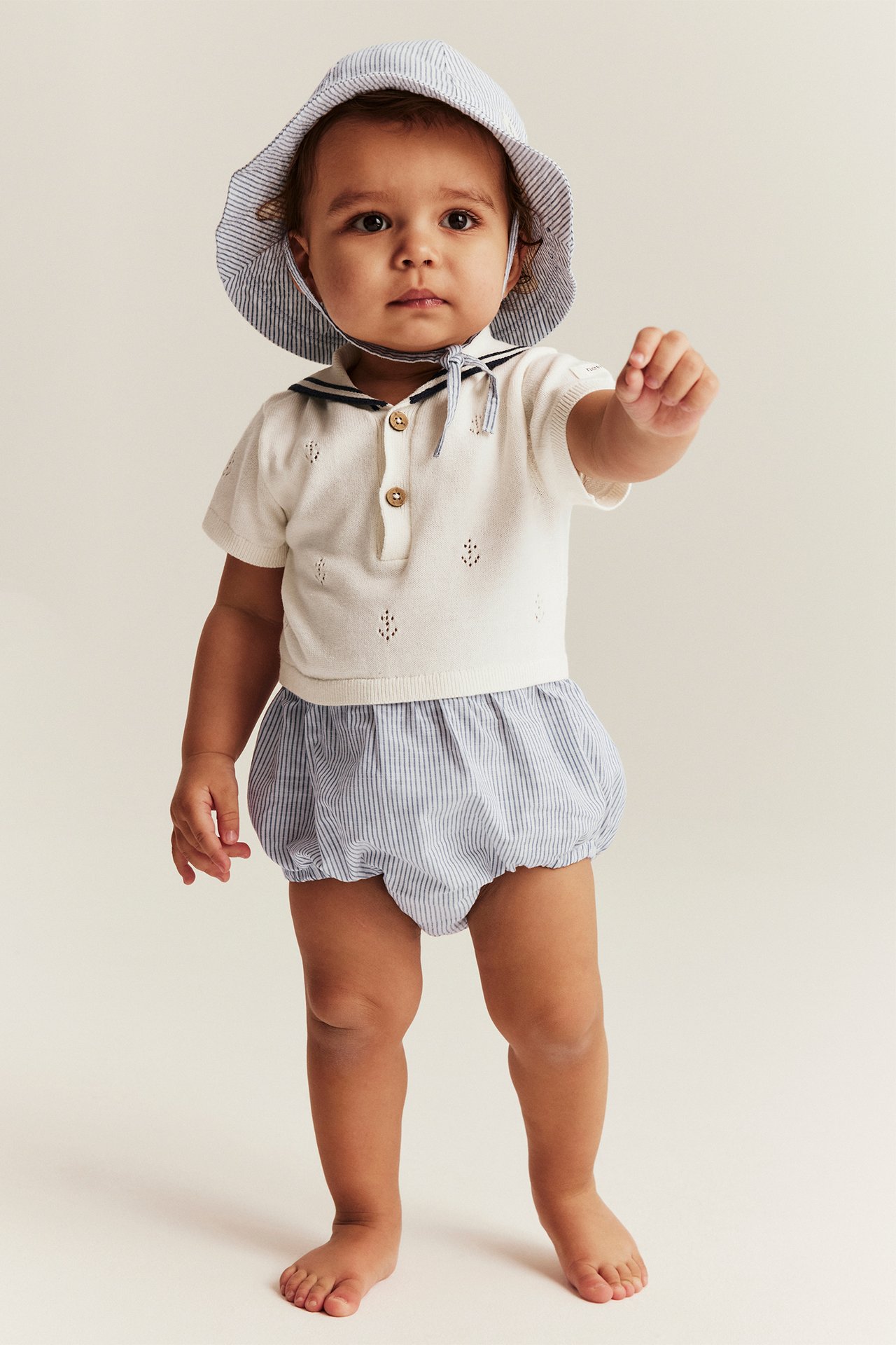 Vorderansicht: Baby in einem cremefarbenen gestrickten Matrosen-Playsuit, gestreiften Bloomers und gestreiftem Sonnenhut.