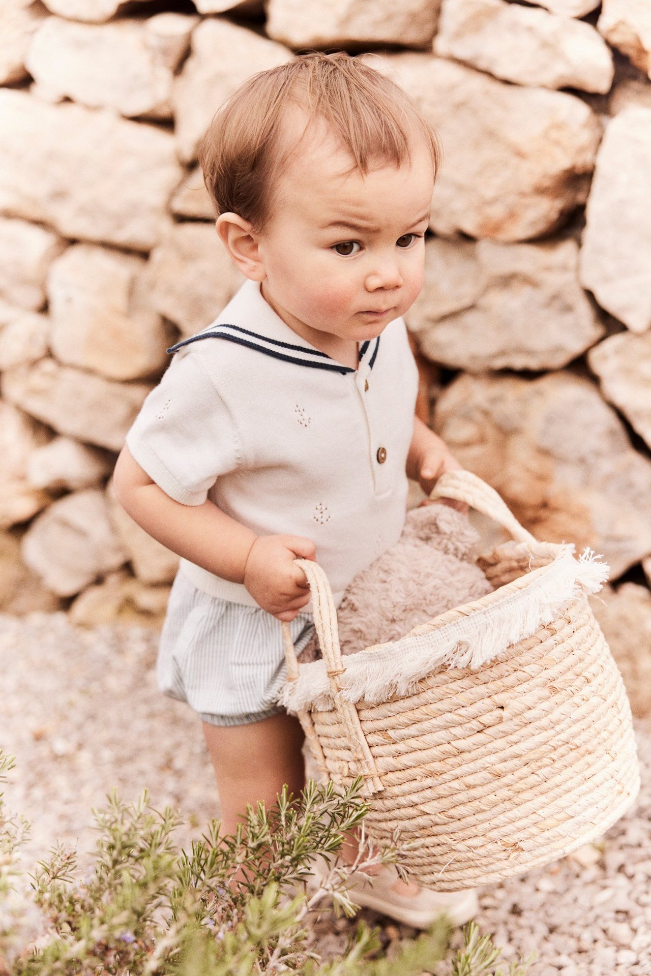 Baby in cremeweißem gestricktem Pointelle-Strampler, Matrosenkragen, gestreiftem Unterteil, hält einen Strohkorb.