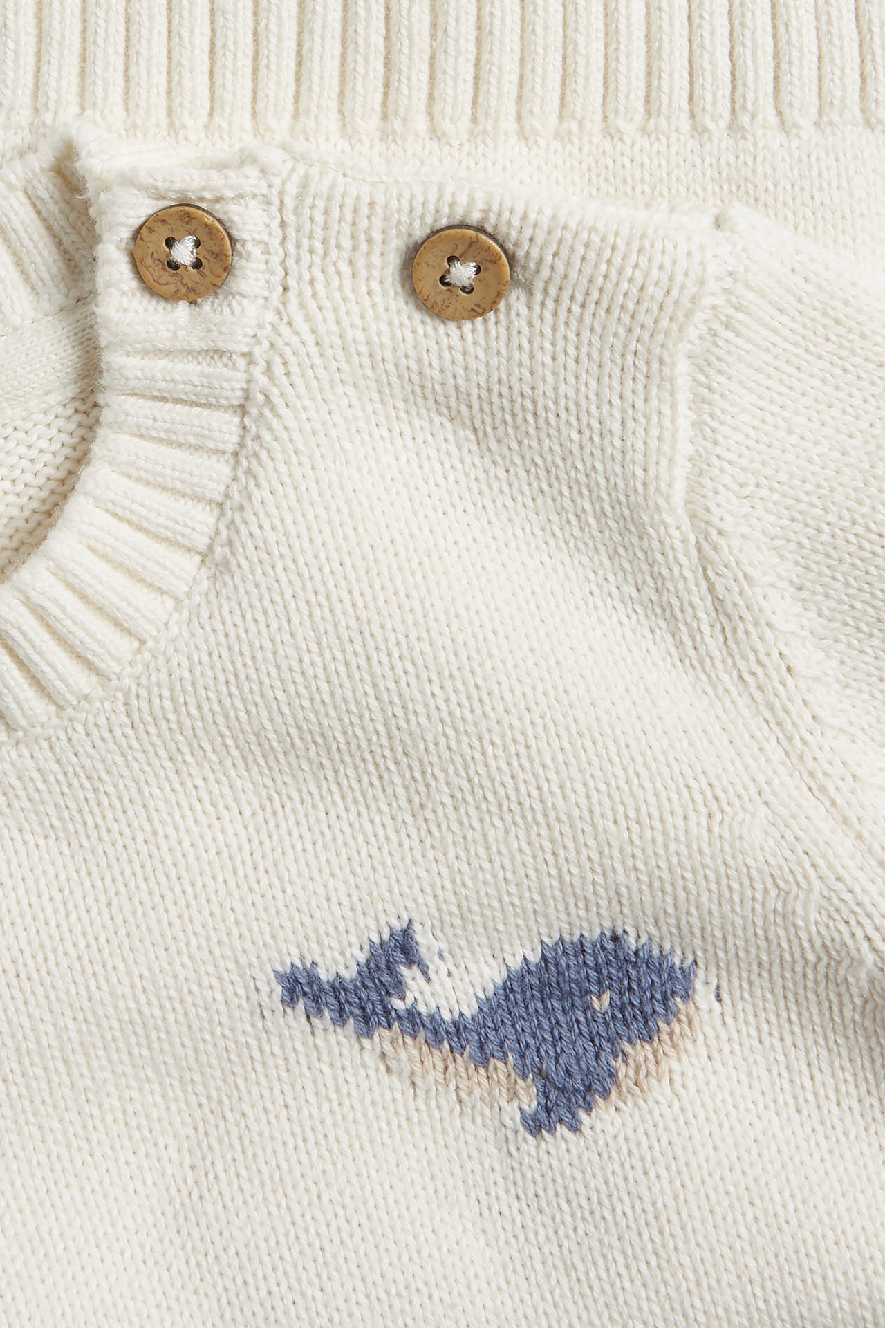 Nahaufnahme eines cremefarbenen gestrickten Baby-Baumwollpullovers mit zwei Holzknöpfen an der Schulter und blauem Walmuster.