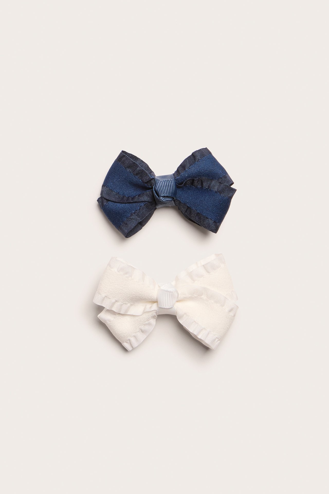 Close-up of two hair clips: a dark blue and an off-white solid bow with ruffled edges.