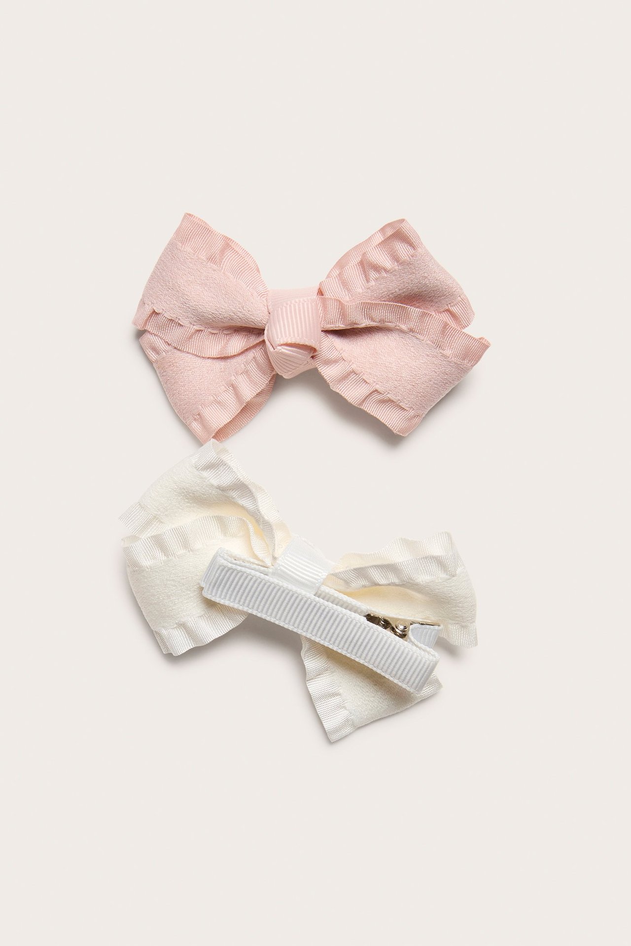 Top-down view of two kid's bow hair clips: light pink and off-white, with frilled edges on metal clips.