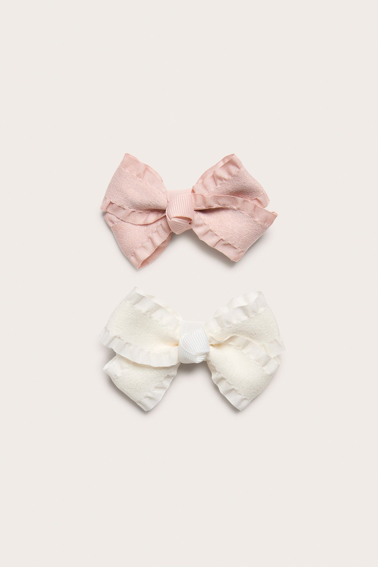 Top view of two kid's hair clips: one light pink, one off-white, both with frilled bows.