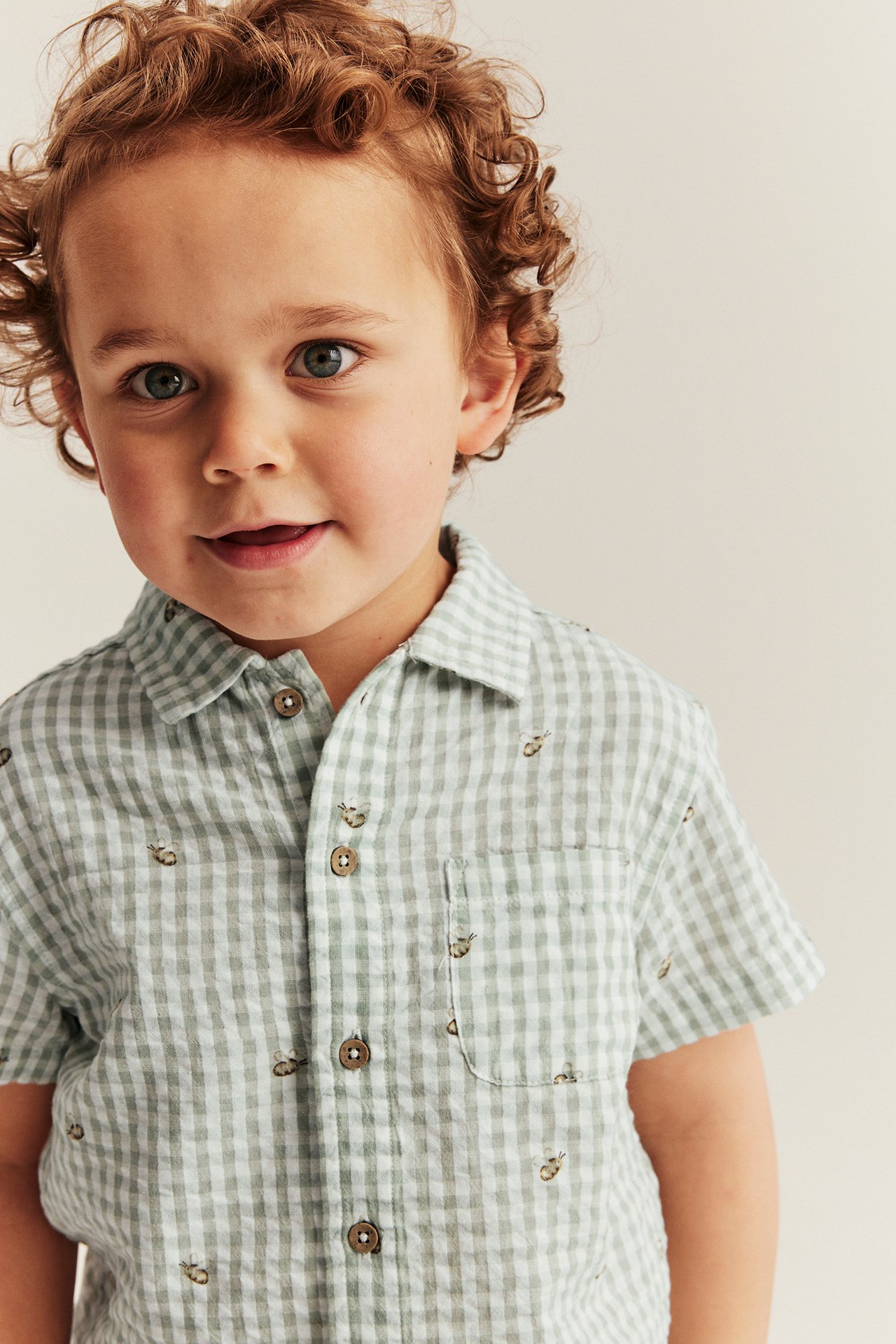 Front view of a kid wearing a light green seersucker short-sleeved shirt with a checked and insect pattern.