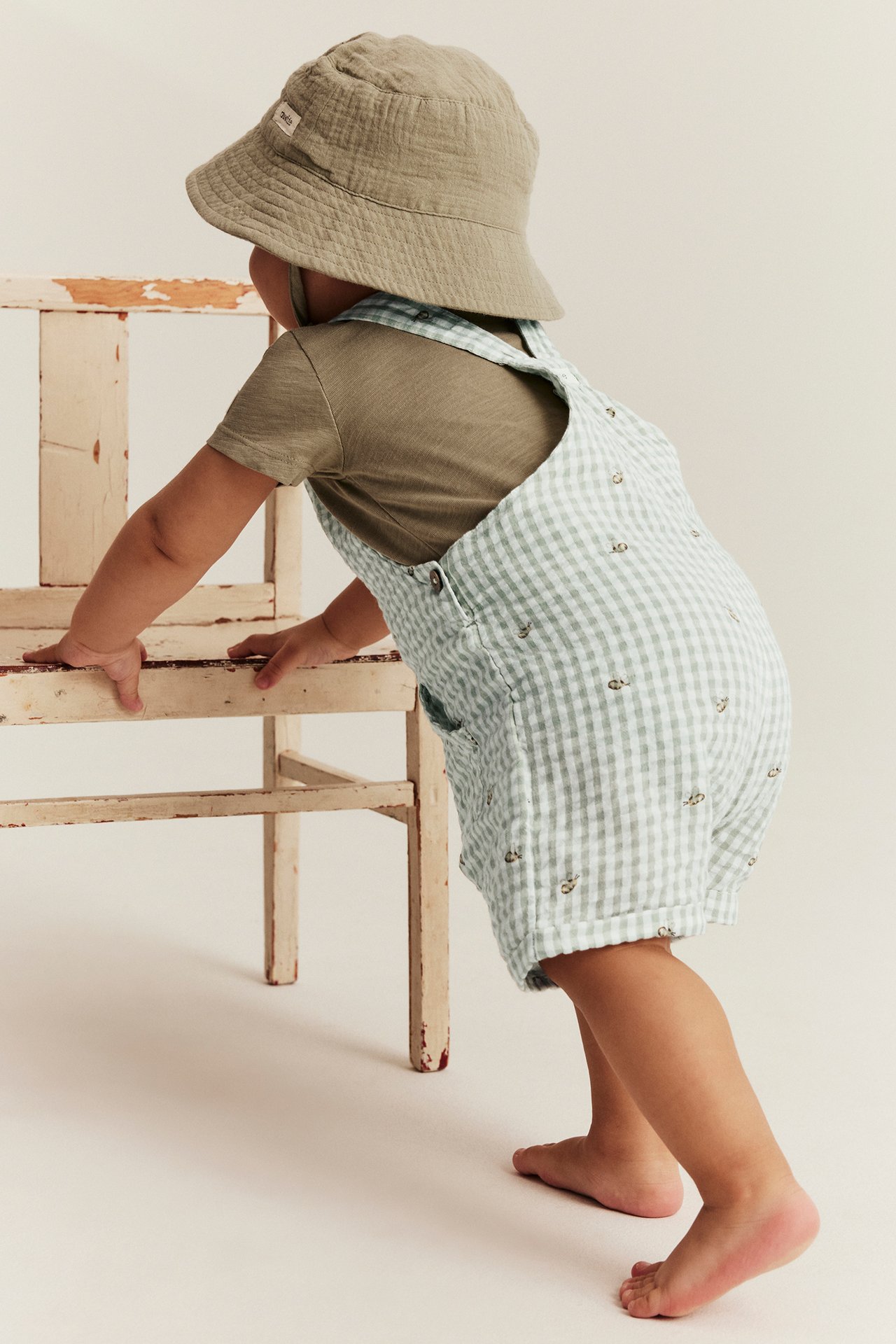 Baby, Rückansicht, in hellgrünen karierten Latzshorts mit Bienenmuster, olivgrünem T-Shirt und beigem Fischerhut.