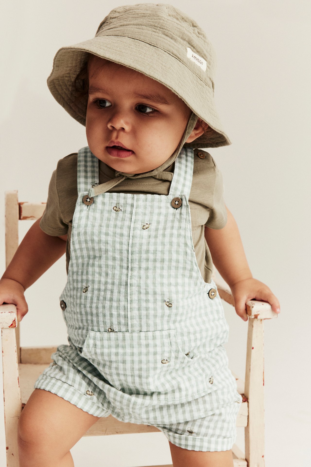 Front view: Baby in light green checked dungaree shorts with bee embroidery, khaki tee, and beige bucket hat.