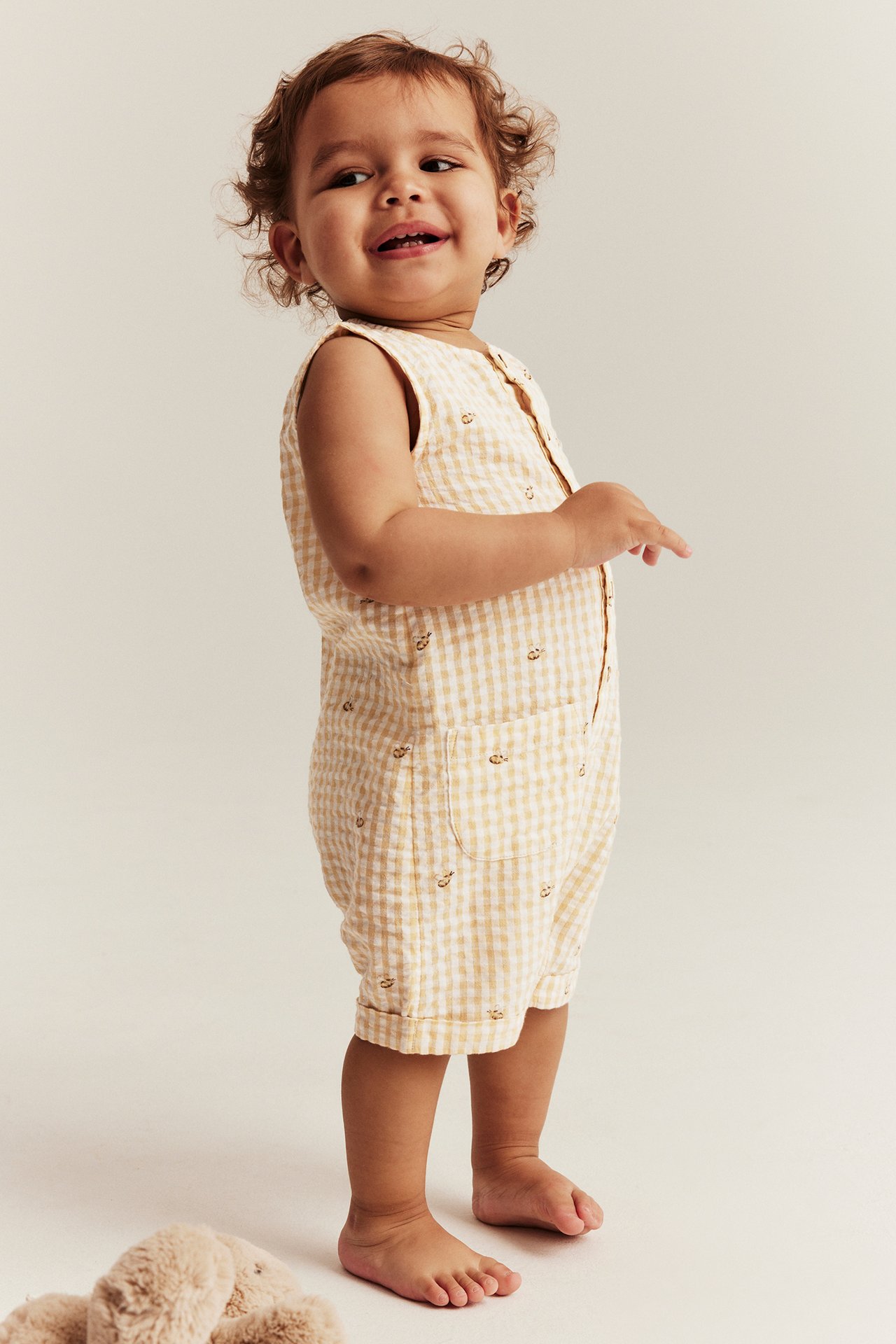 Front view: Baby in a yellow and white gingham seersucker playsuit with animal motifs and front buttons.