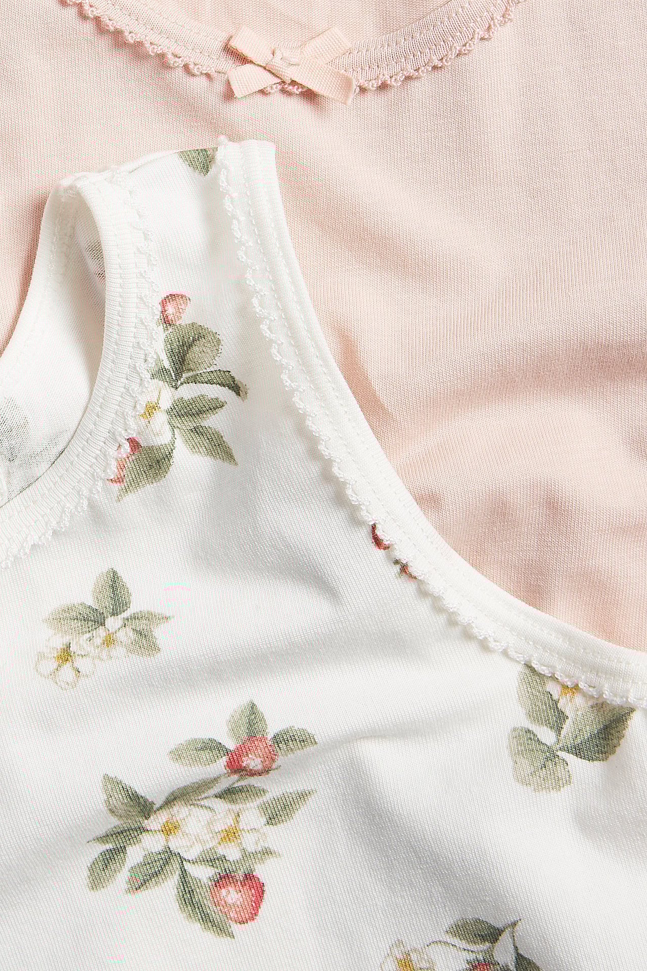 Nahaufnahme: Off-weißes geblümtes Baby-Tanktop mit Wellenkanten, daneben ein rosa Tanktop mit Schleife.