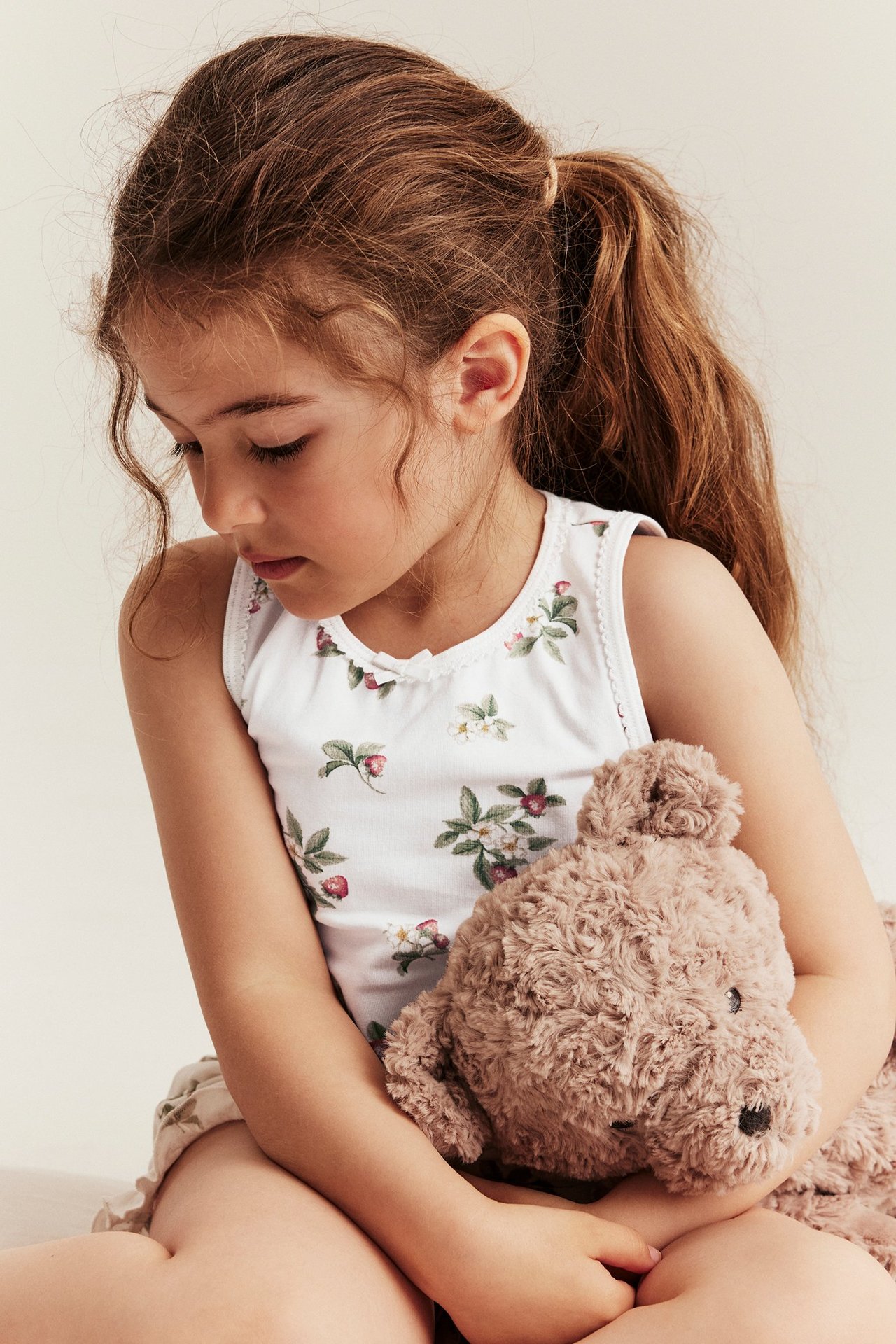 Vorderansicht eines Kindes in cremeweißem geblümten Tanktop mit Wellenkanten und Schleifen, hält braunen Teddybär.