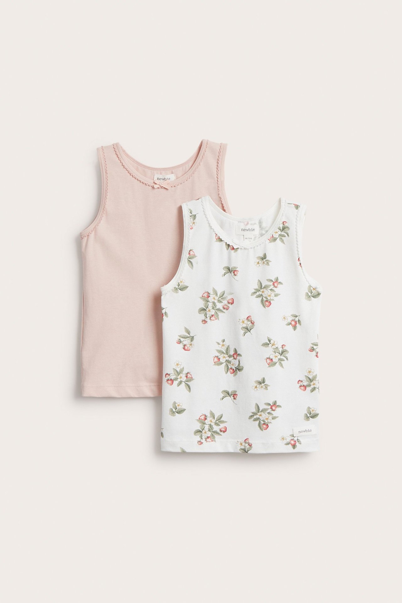 Vorderansicht: zwei Kinder-Baumwoll-Tanktops. Cremeweißes Blumenmuster und einfarbiges Hellrosa, mit Wellenkanten und kleinen Schleifen.