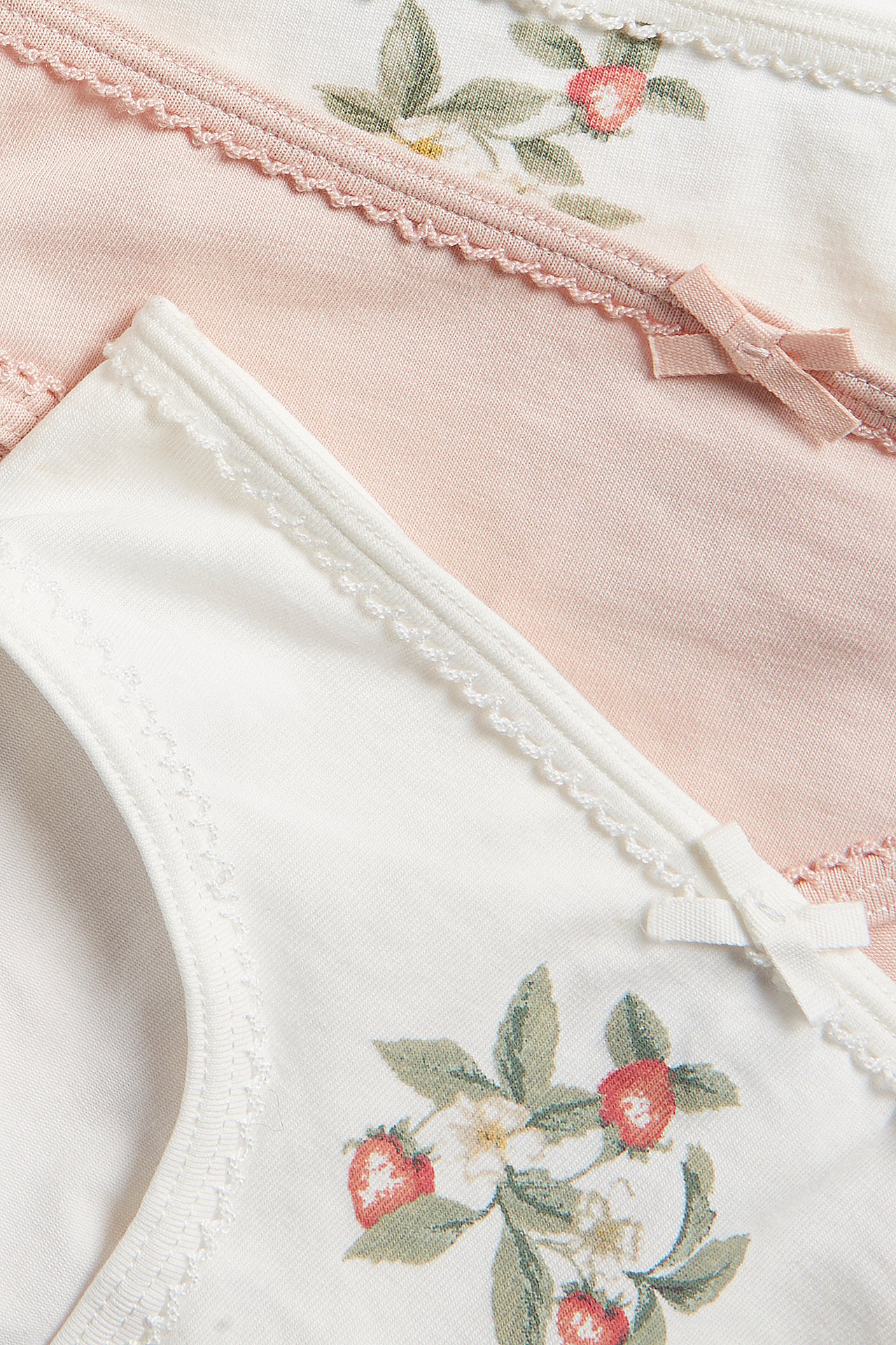 Close-up of three jersey panties: two off-white with strawberry print, one light pink, with bows and scalloped edges.