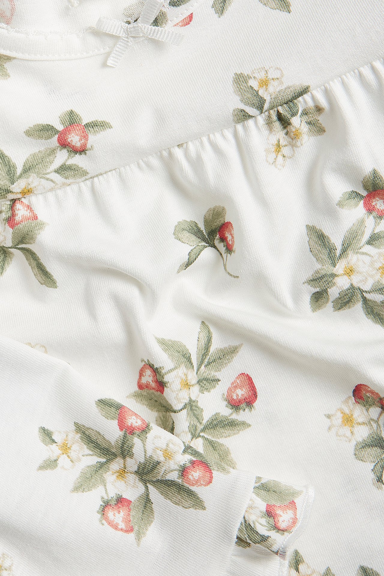 Nahaufnahme von cremeweißem Baumwollpyjama mit Erdbeermuster, kleiner Schleife und Rüschen.