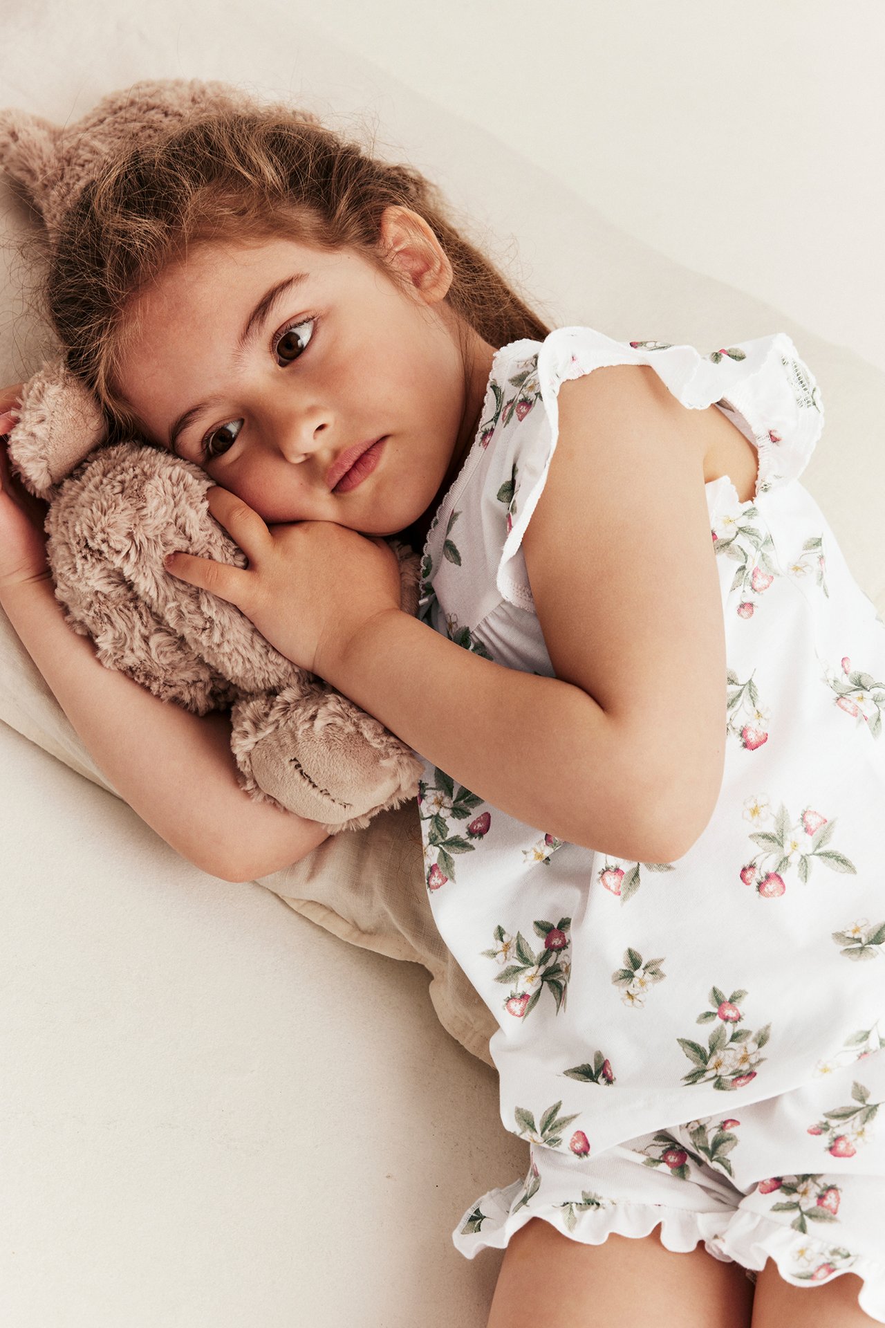 Draufsicht eines Kindes in einem cremefarbenen Erdbeermuster-Pyjama mit Rüschen, das einen braunen Teddybär hält.