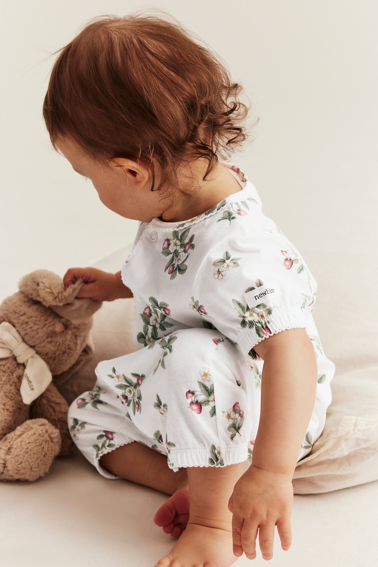 Seitenansicht eines Babys in cremeweißem Baumwoll-Strampler mit Erdbeermuster, Picot-Kanten, das einen braunen Hasen berührt.