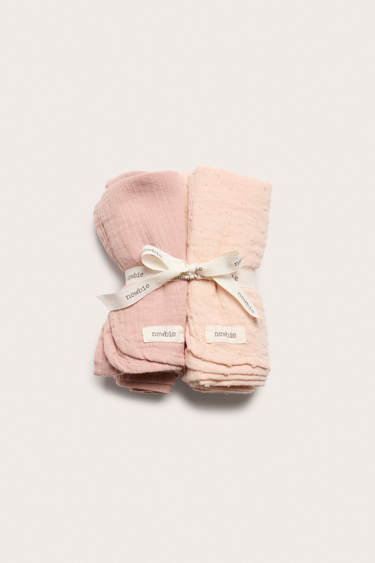 Top-down view: Two folded cotton muslin blankets, one solid pink, one light beige patterned, tied with ribbon.