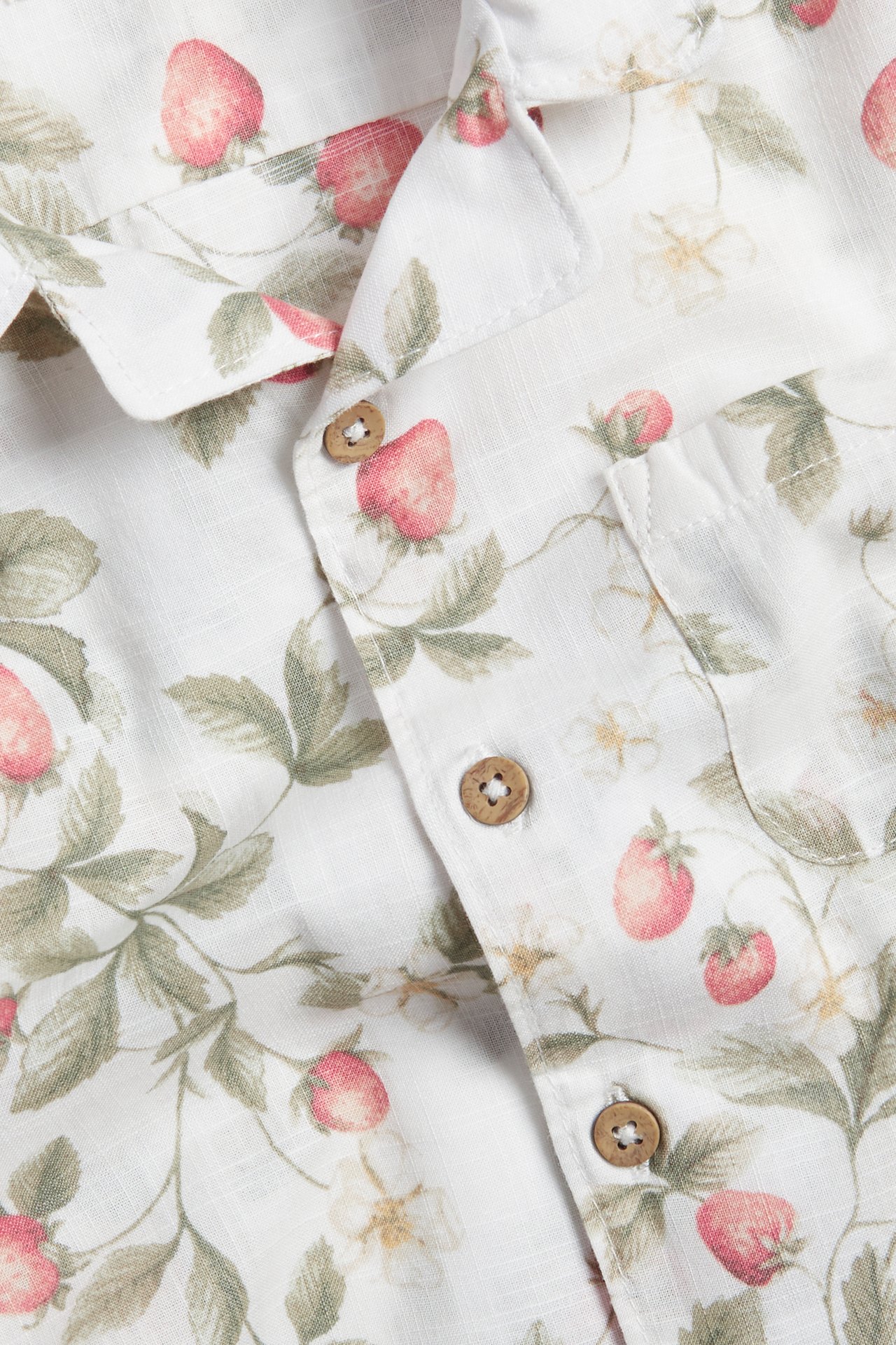 Close-up front view: off-white cotton shirt with red strawberry pattern, wooden buttons, and chest pocket.