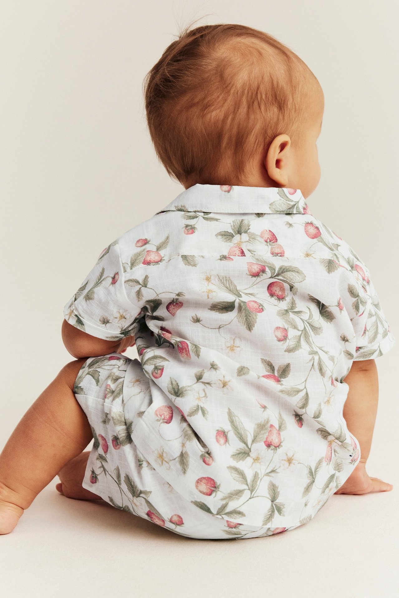 Rückenansicht eines Babys in einem cremeweißen Baumwoll-Kurzarmhemd und passenden Shorts mit Erdbeermuster.