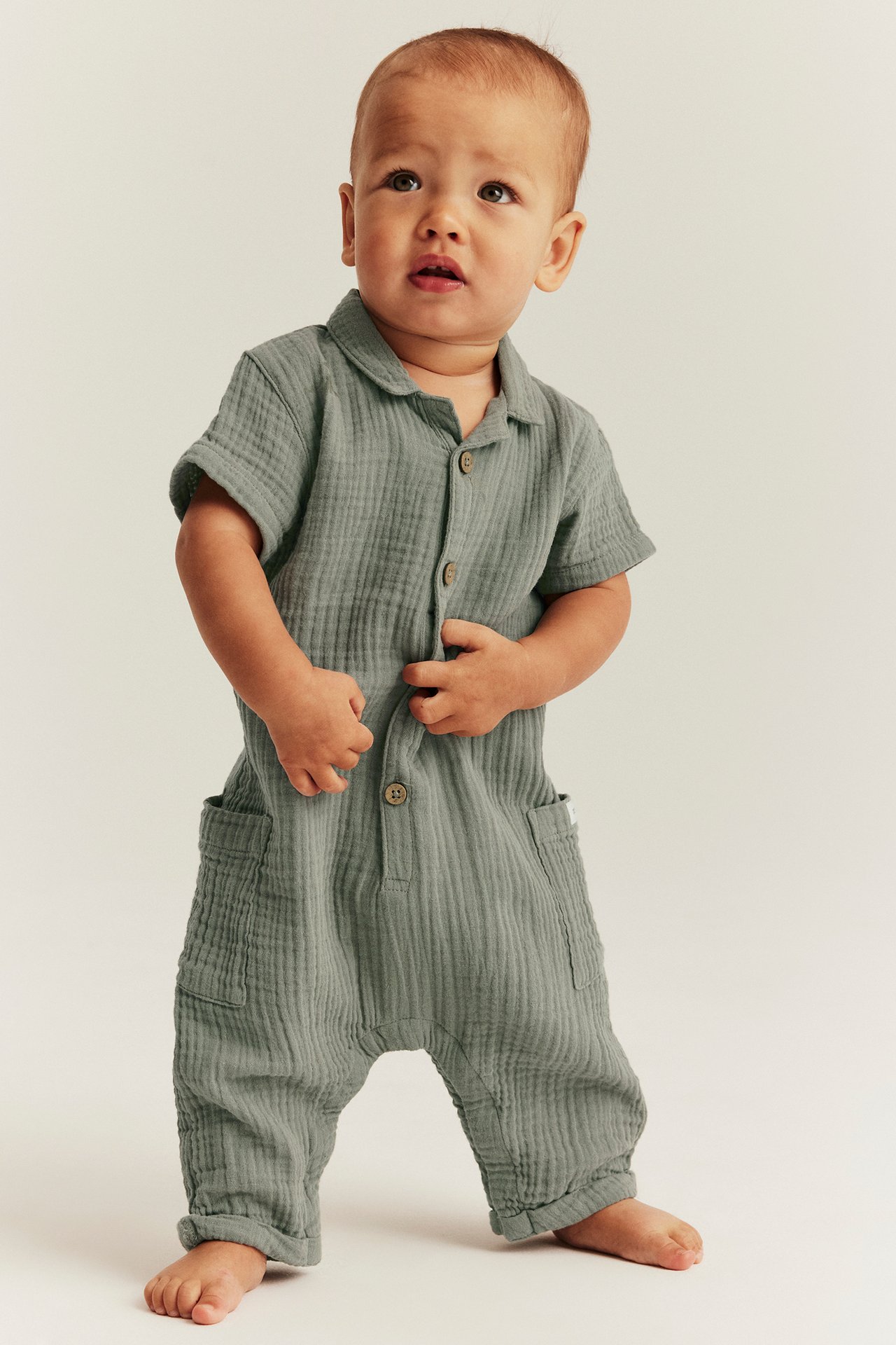 Vorderansicht eines Babys in grünem, kurzärmeligem Baumwoll-Playsuit mit Kragen, Knöpfen und Seitentaschen.