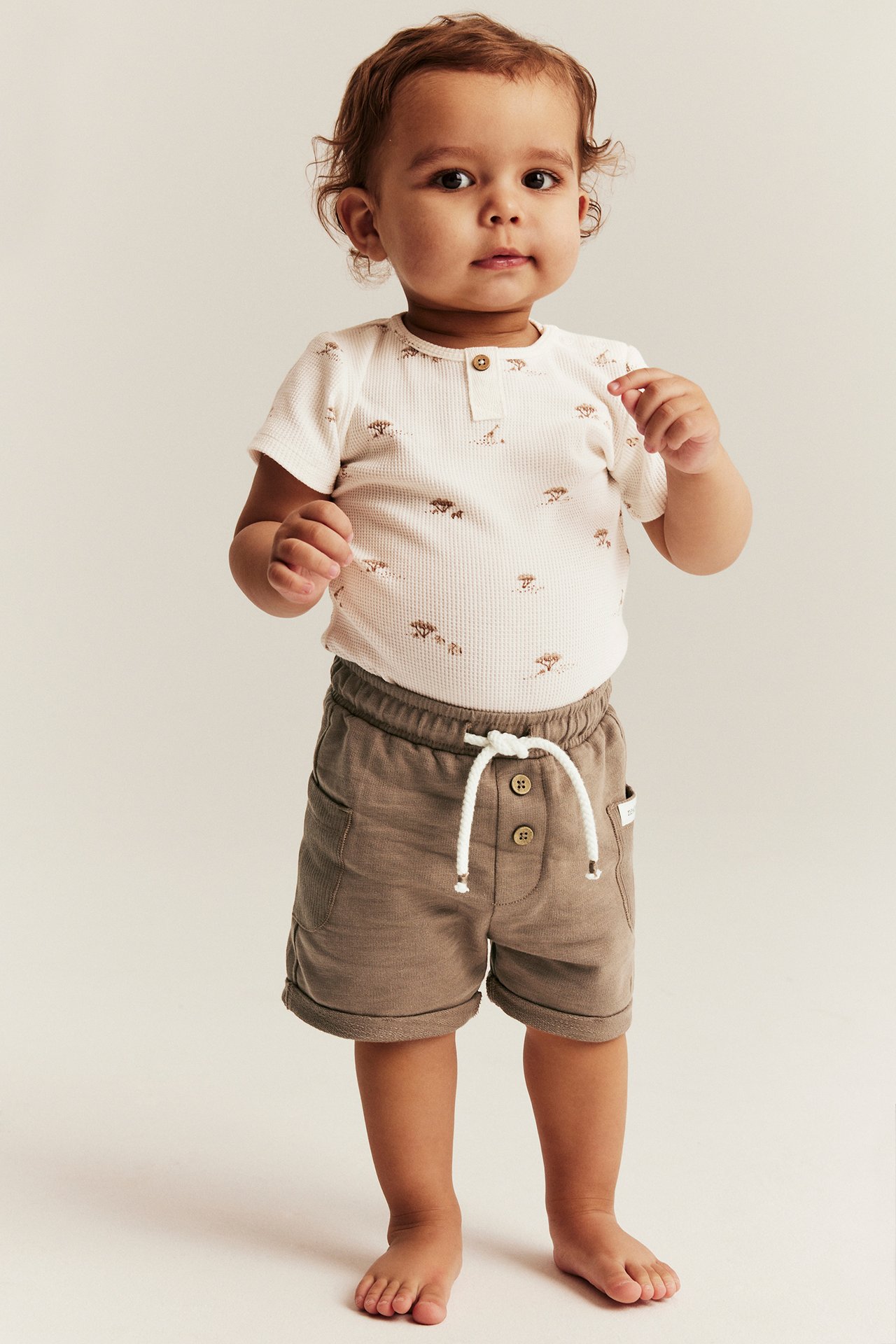 Front view of a baby wearing brown cotton shorts with a white drawstring and a white patterned short-sleeved bodysuit.