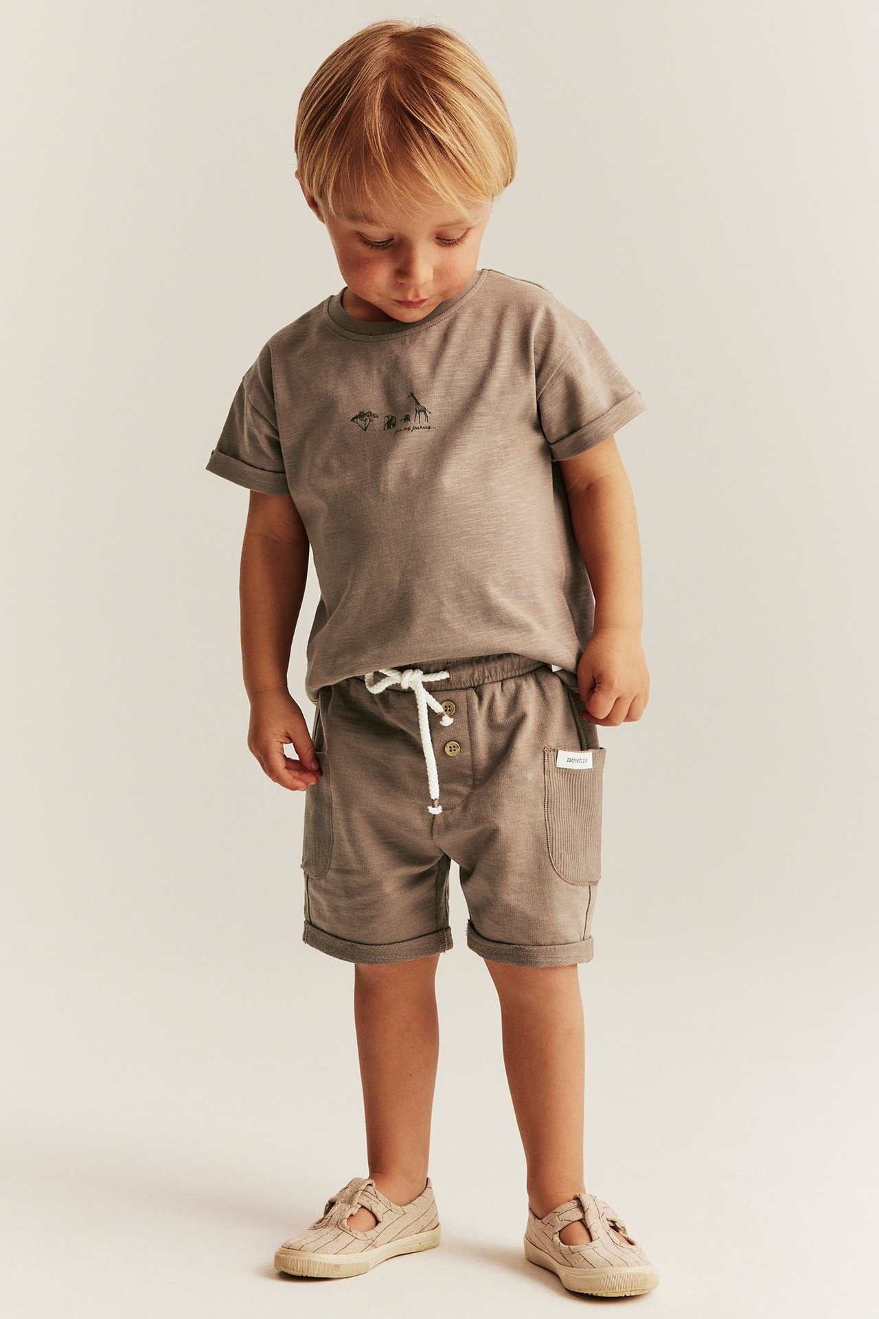 Vorderansicht eines Kindes in braunen Slub-Jersey-Shorts mit Taschen, einem braunen T-Shirt und beigen Sandalen.