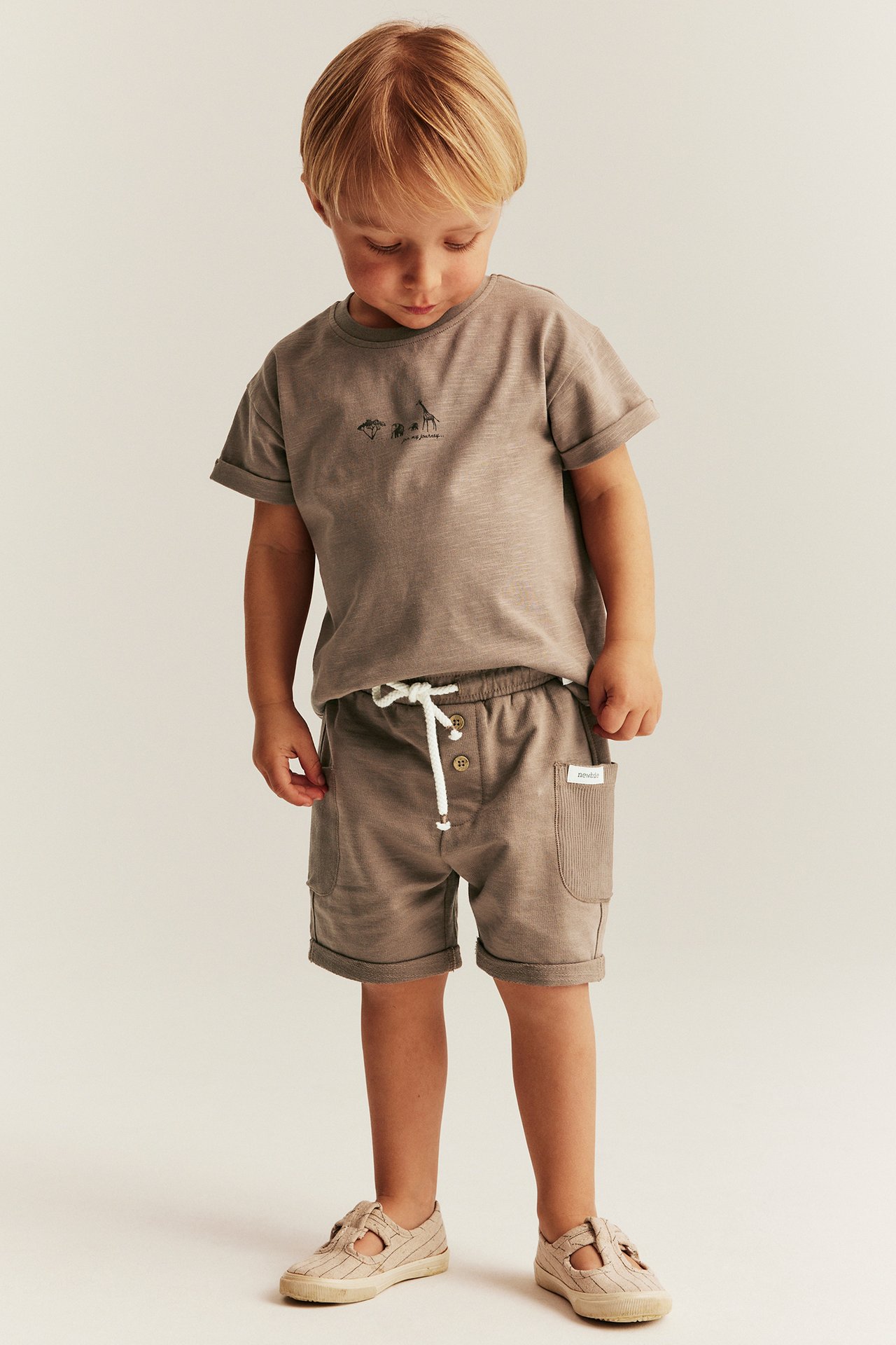Vorderansicht eines Kindes in braunen Slub-Jersey-Shorts mit Taschen, einem braunen T-Shirt und beigen Sandalen.