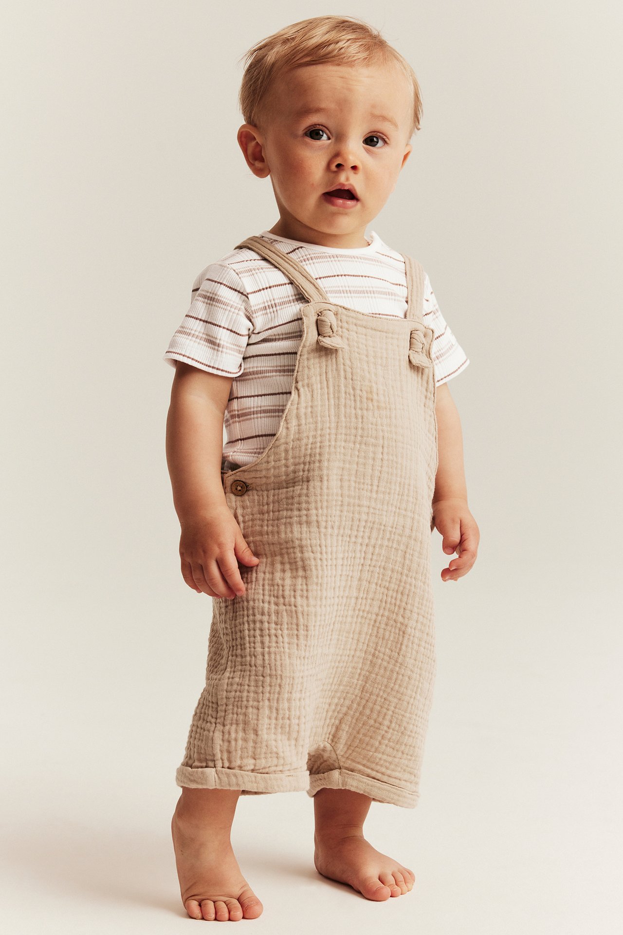 Vorderansicht eines Babys in beigem strukturiertem Overall über einem weiß gestreiften kurzärmeligen Body.