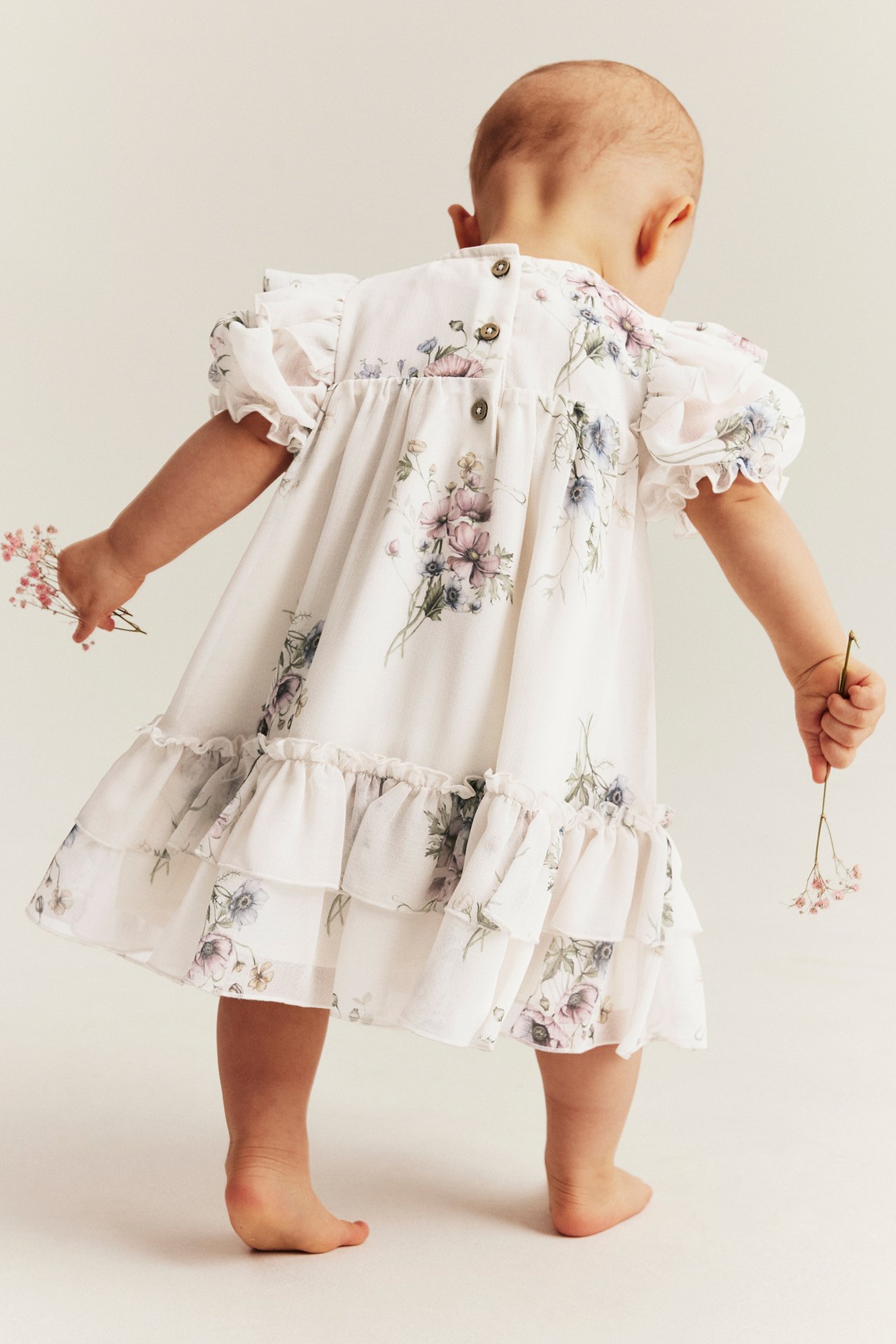 Back view: baby in an off-white floral chiffon dress with puff sleeves, ruffles, holding small flowers.