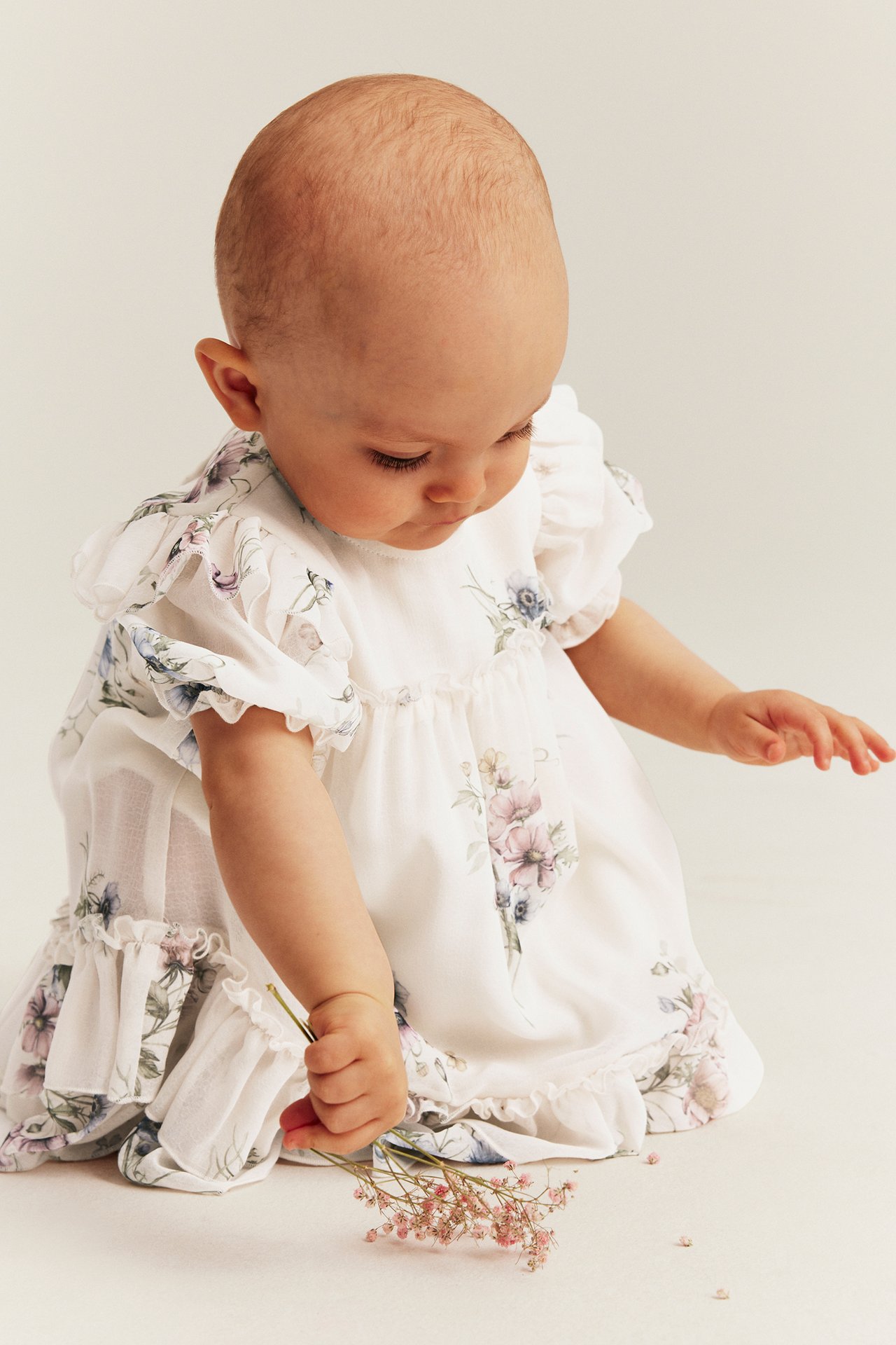 Front view of a baby wearing an off-white floral chiffon dress with puff sleeves, holding small pink flowers.