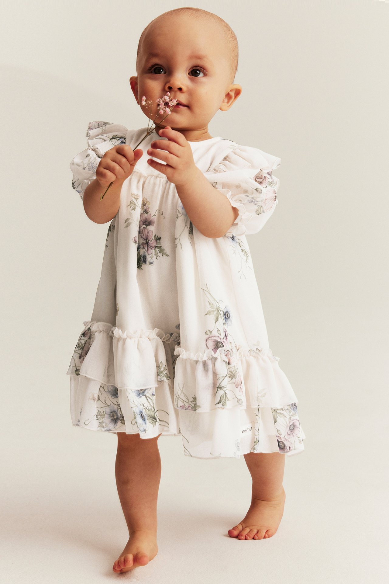 Front view: Baby in off-white floral chiffon dress with puff sleeves, ruffled skirt, holding pink flower.