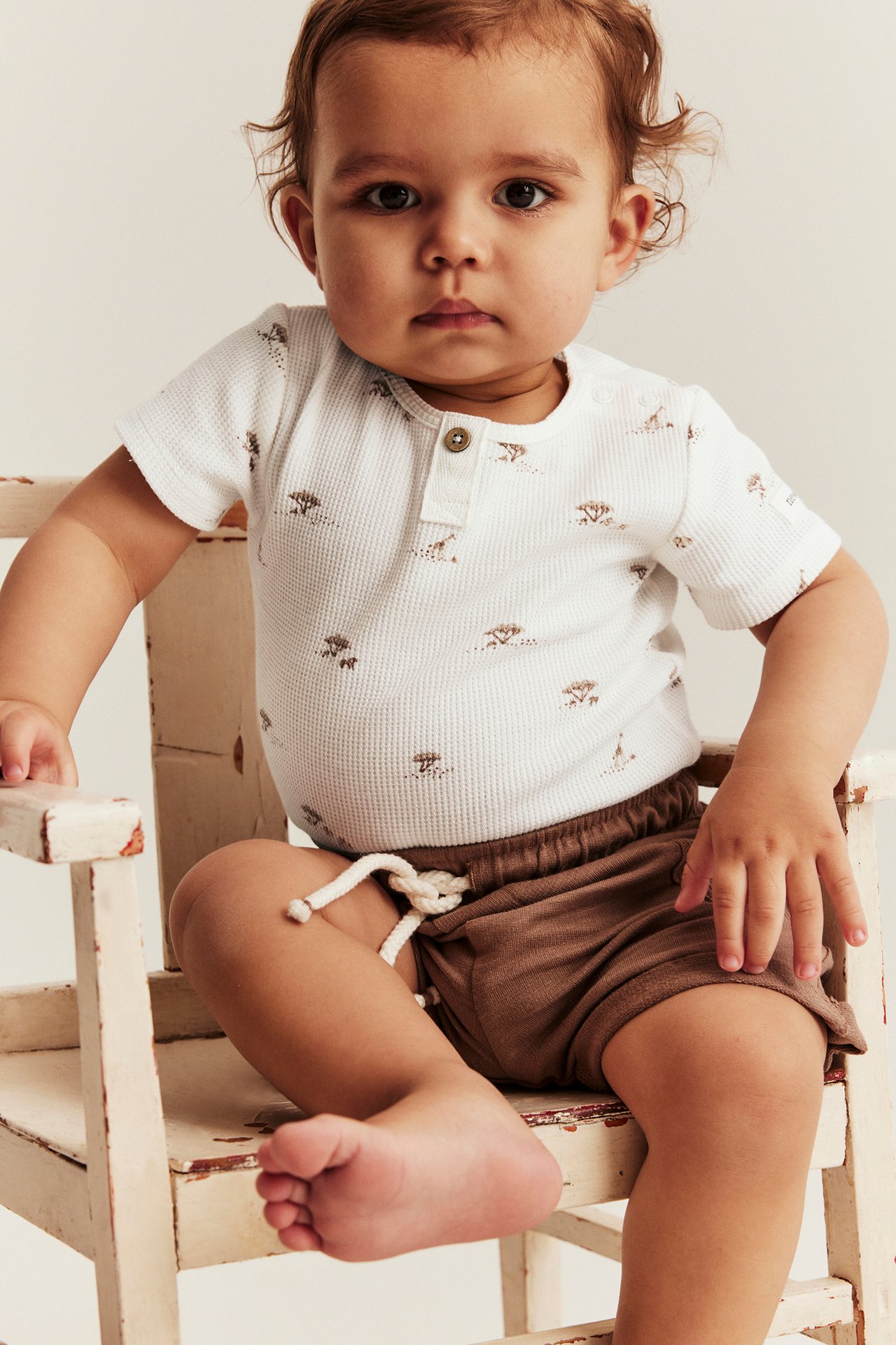 Vorderansicht eines Babys in einem cremefarbenen, waffelgestrickten Kurzarm-Body mit Tiermuster und braunen Shorts.