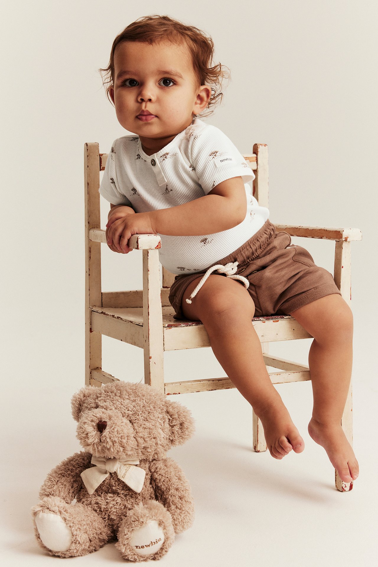 Vorderansicht eines Babys in einem cremefarbenen, tiermuster-Waffelstrick-Body, braunen Shorts, neben einem Teddybär.