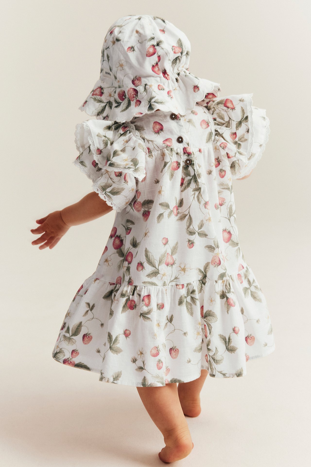 Back view of a baby wearing an offwhite cotton strawberry-patterned dress with ruffled sleeves and a matching hat.
