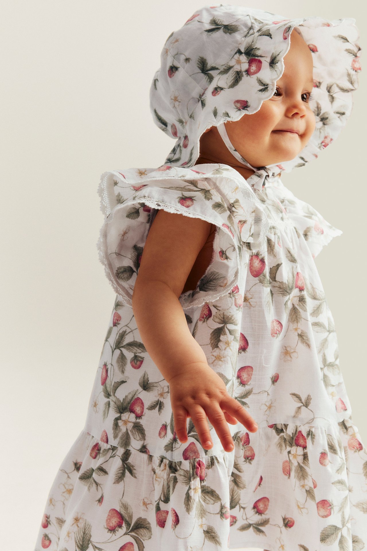 Side view of a baby wearing an offwhite cotton strawberry-patterned dress with butterfly sleeves and a matching bonnet.