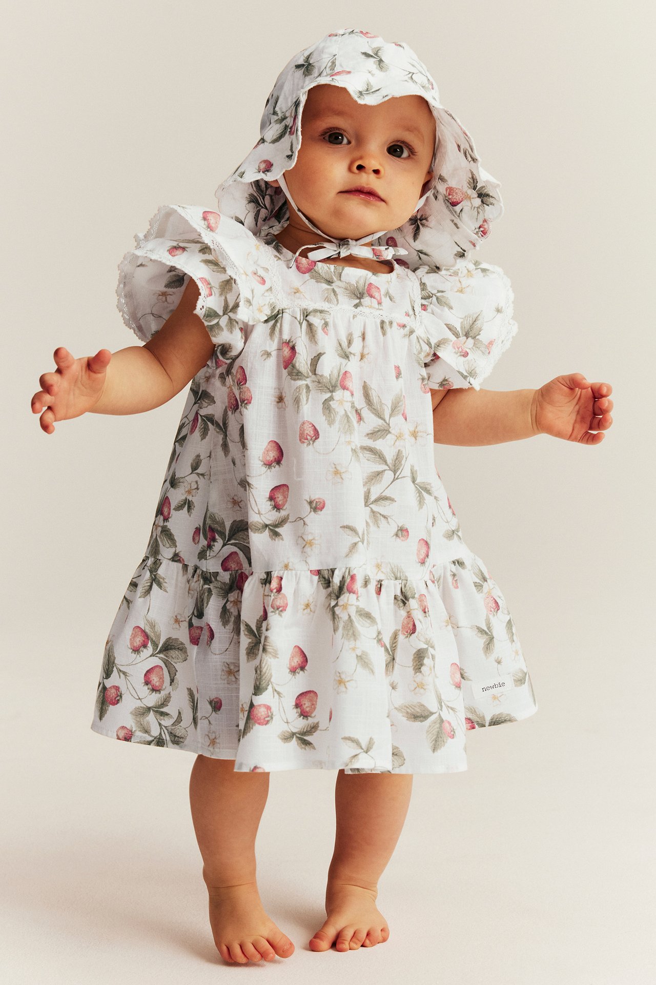 Front view: baby in off-white cotton strawberry-patterned dress with butterfly sleeves and matching sun hat.
