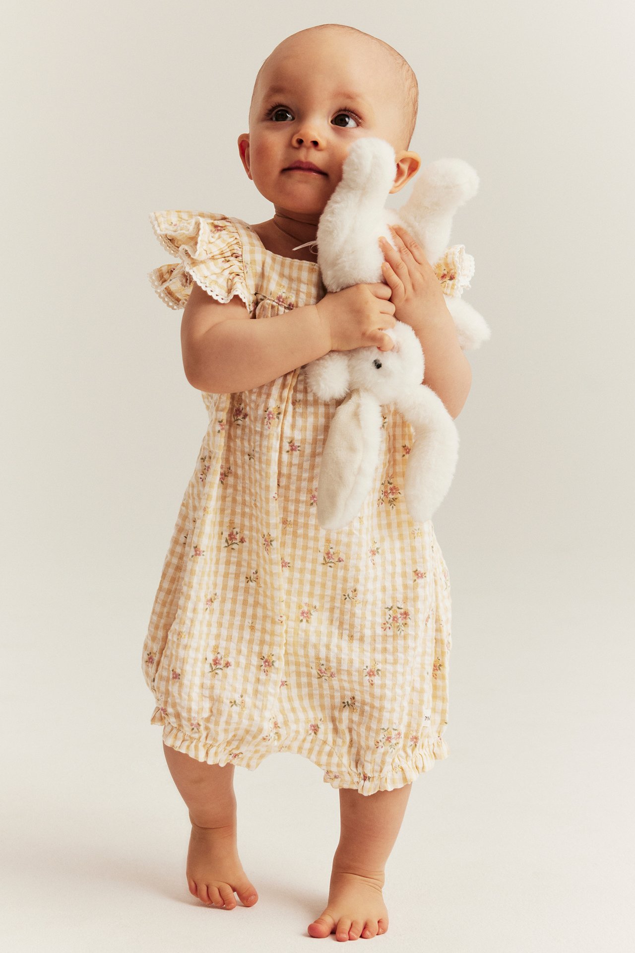 Front view: baby in a yellow gingham floral cotton playsuit with ruffle sleeves, holding a white plush bunny.