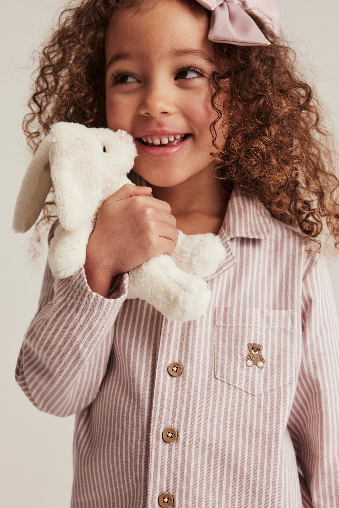 Nahaufnahme eines Kindes in einem rosa gestreiften Baumwoll-Pyjamahemd mit gesticktem Bären, hält ein weißes Häschen.