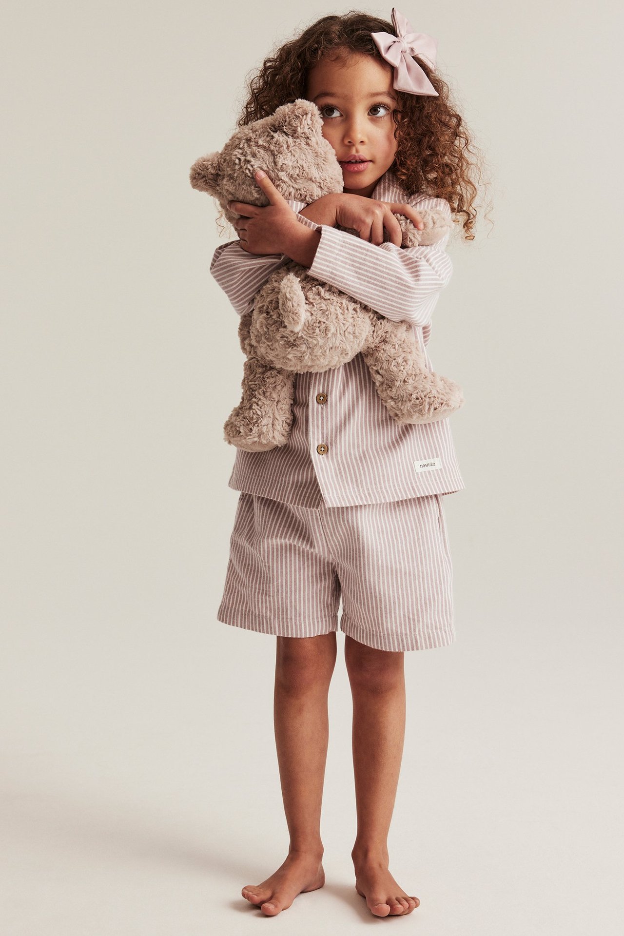 Ganzansicht eines Kindes in rosa gestreiftem Pyjama, das einen braunen Teddybär hält, mit rosa Schleife.