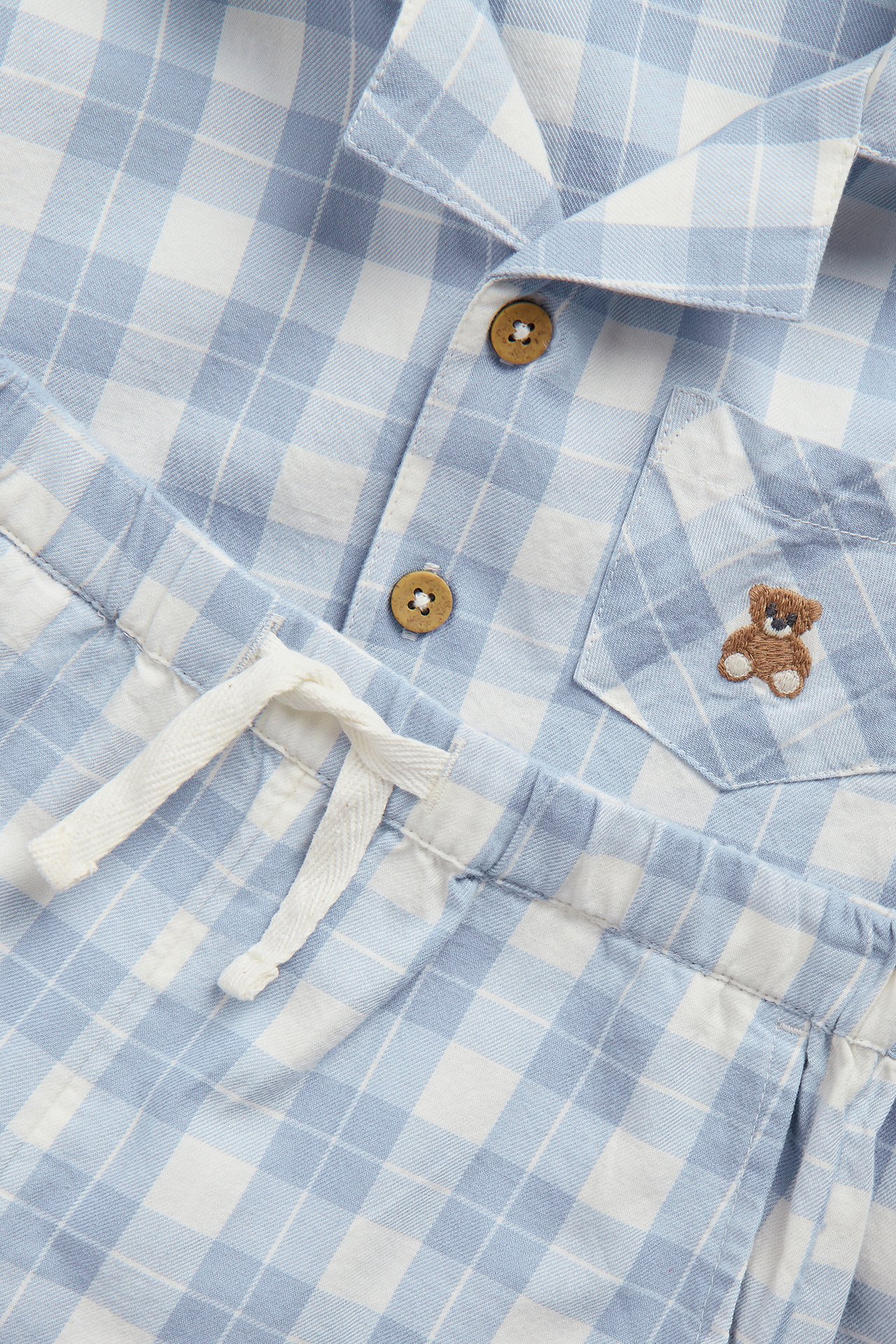 Nahaufnahme eines blau-weiß karierten Pyjama-Sets. Hemd mit Knöpfen, Teddybär-Tasche und Shorts mit Kordelzug.
