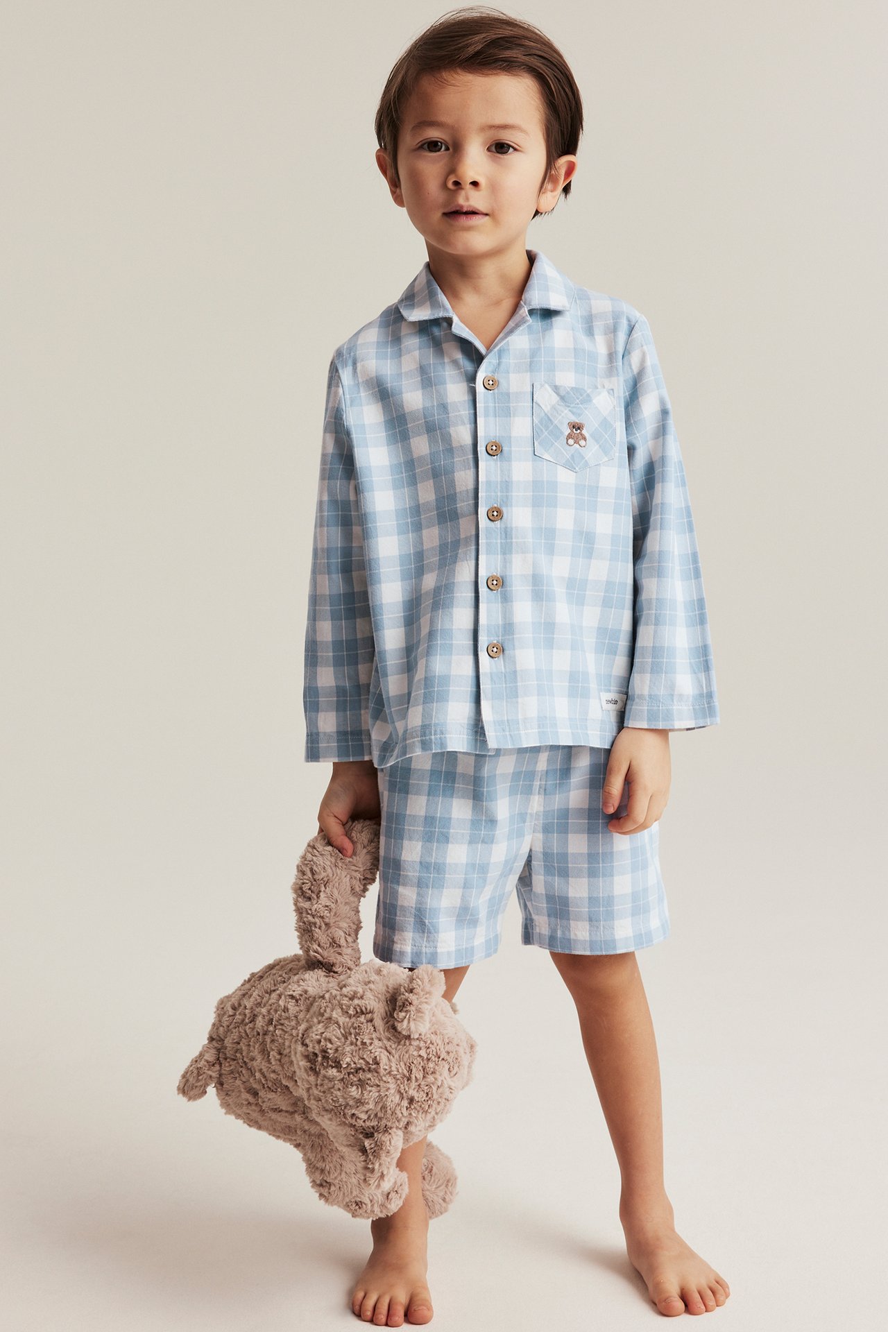 Vorderansicht eines Kindes in blau-weiß kariertem Baumwollpyjama, hält einen braunen flauschigen Teddybär.